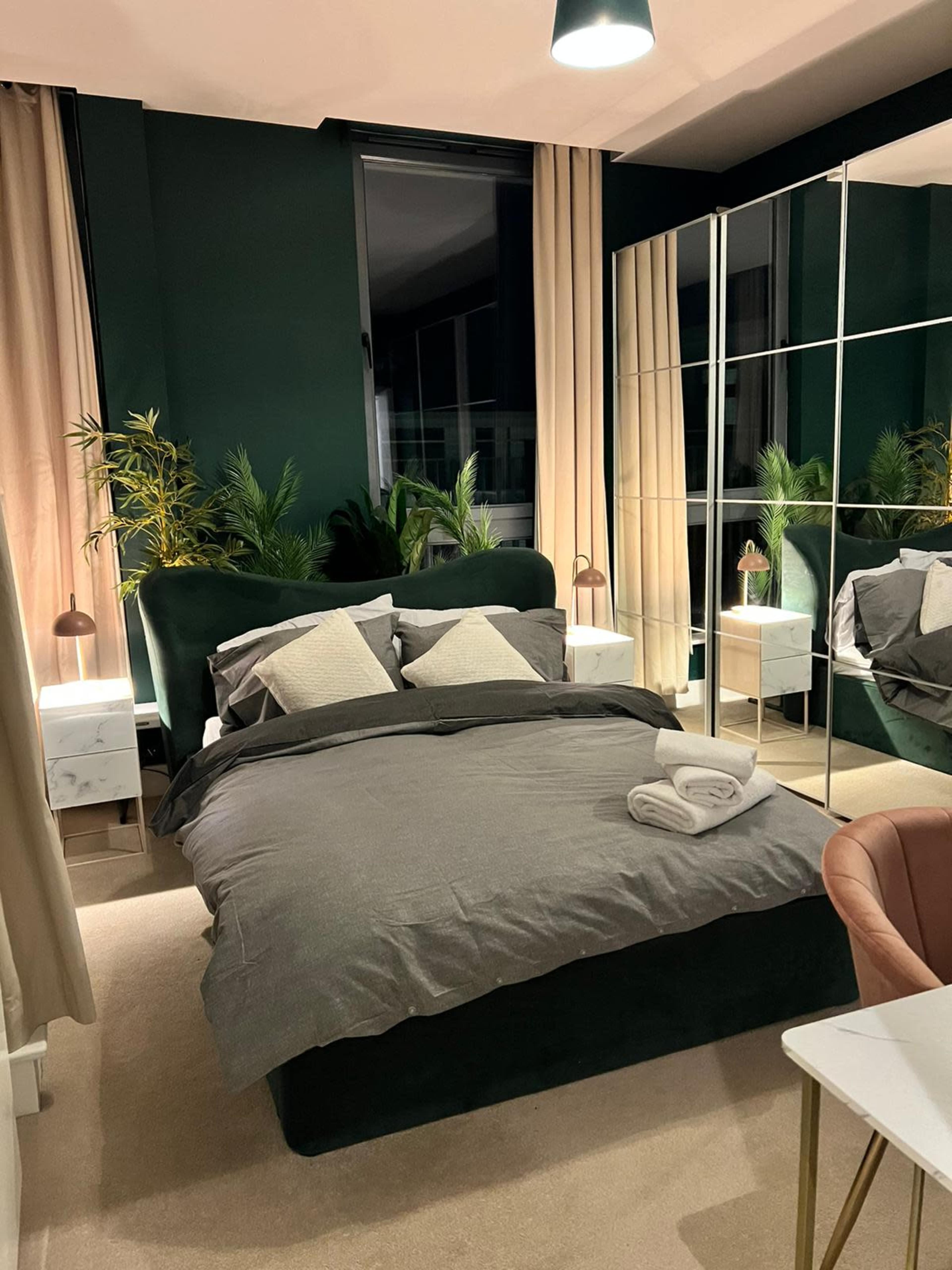 A modern bedroom with a dark green accent wall, a large bed covered in gray bedding, and a mirrored wardrobe beside potted plants and nightstands.