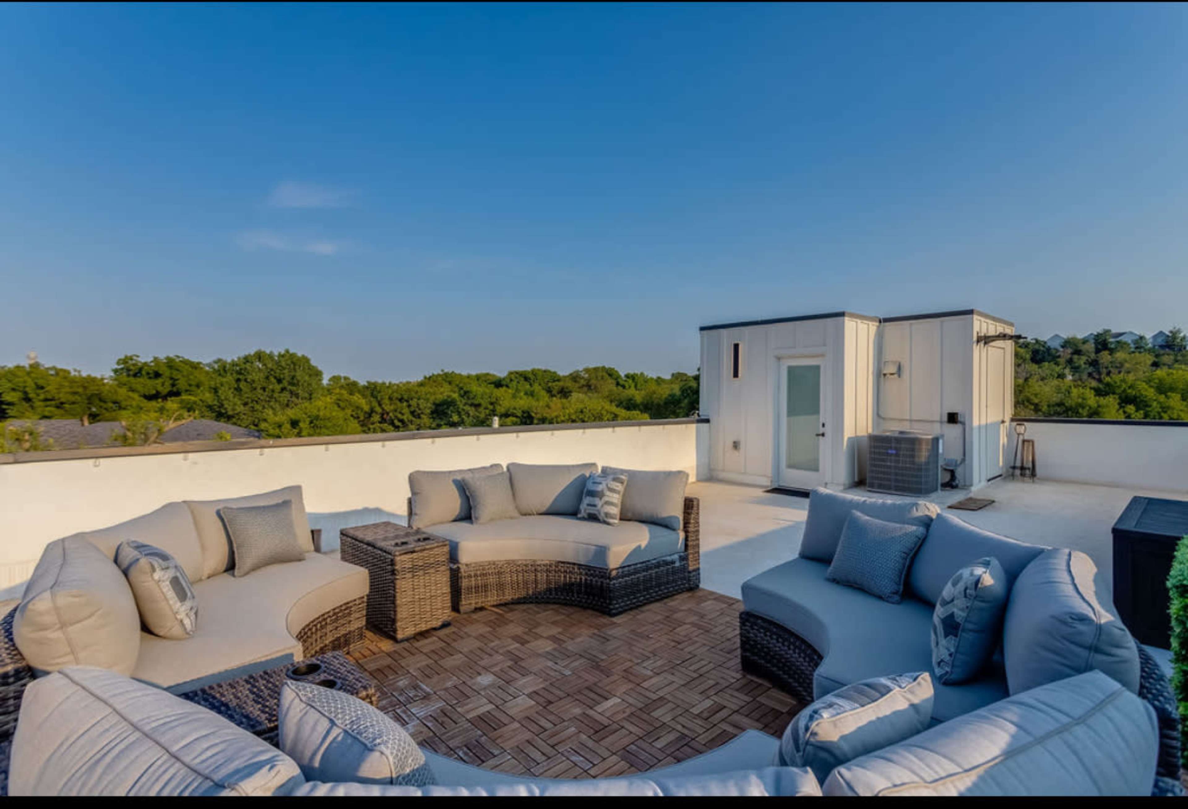 The image shows a rooftop patio with stylish wicker furniture arranged in a circular pattern, overlooking a green landscape.