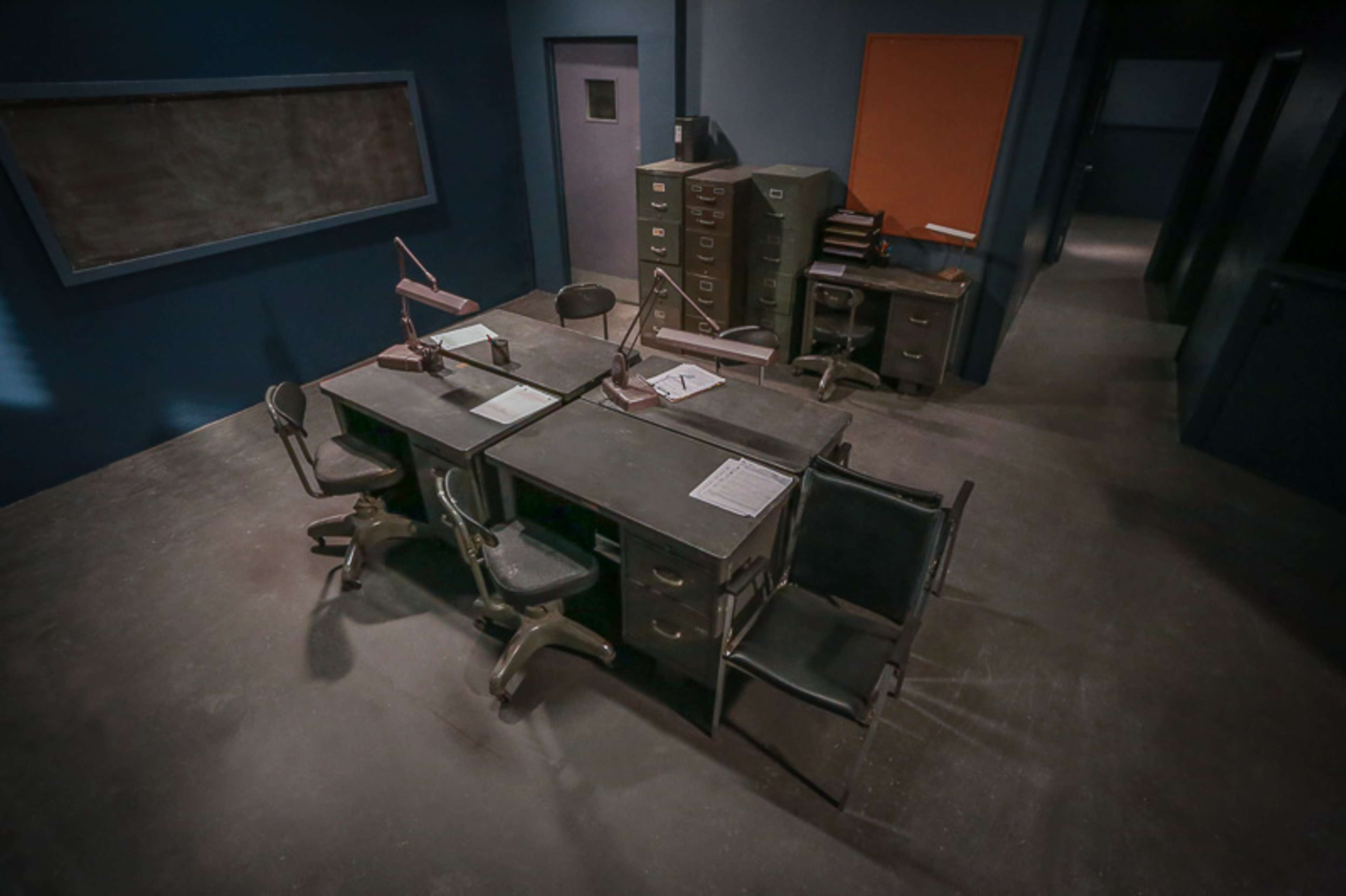 A sparse, dimly lit room featuring a central table surrounded by chairs and several filing cabinets against the walls.