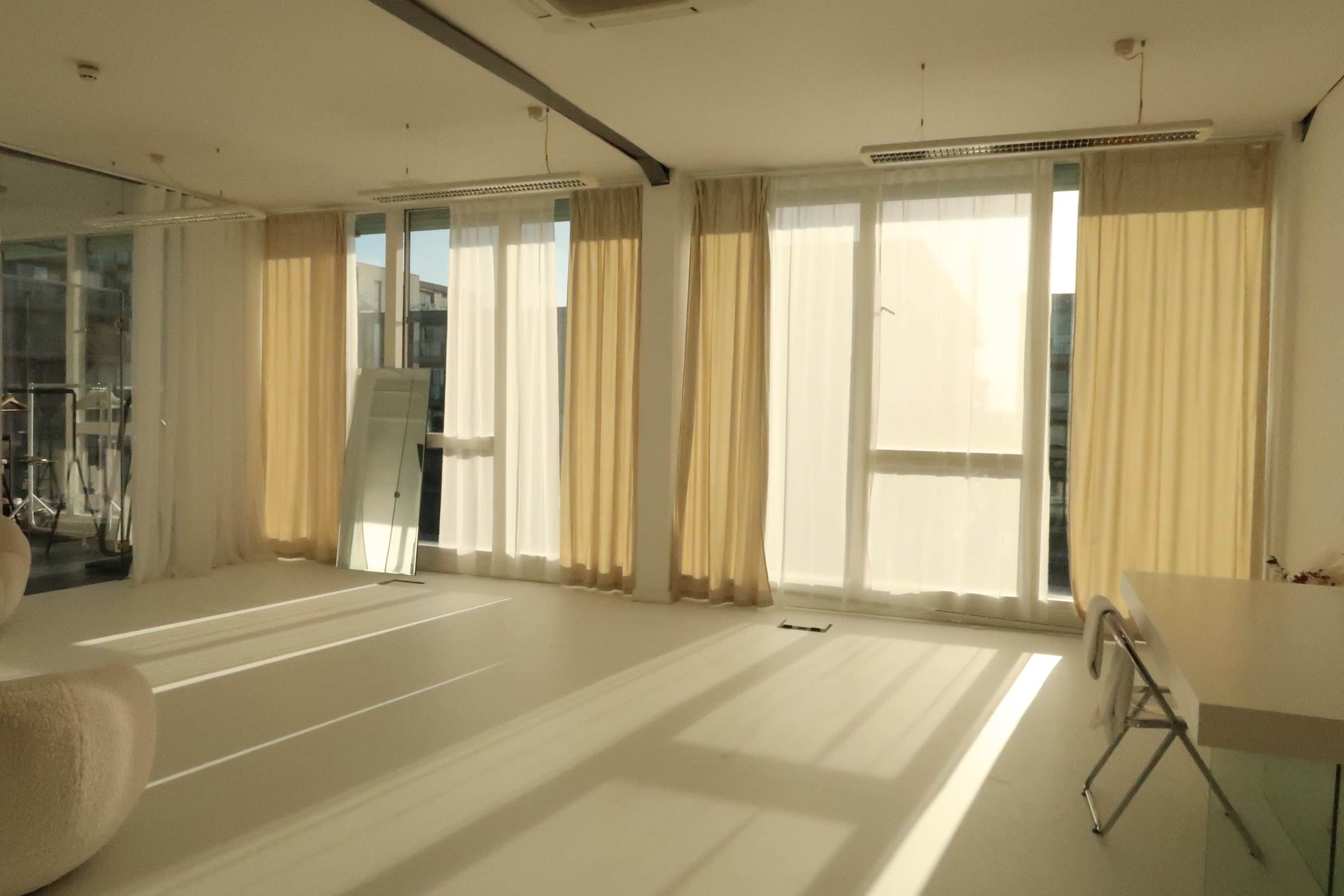 The image shows a bright, spacious room with large windows draped in light curtains, and sunlight casting shadows on the floor.