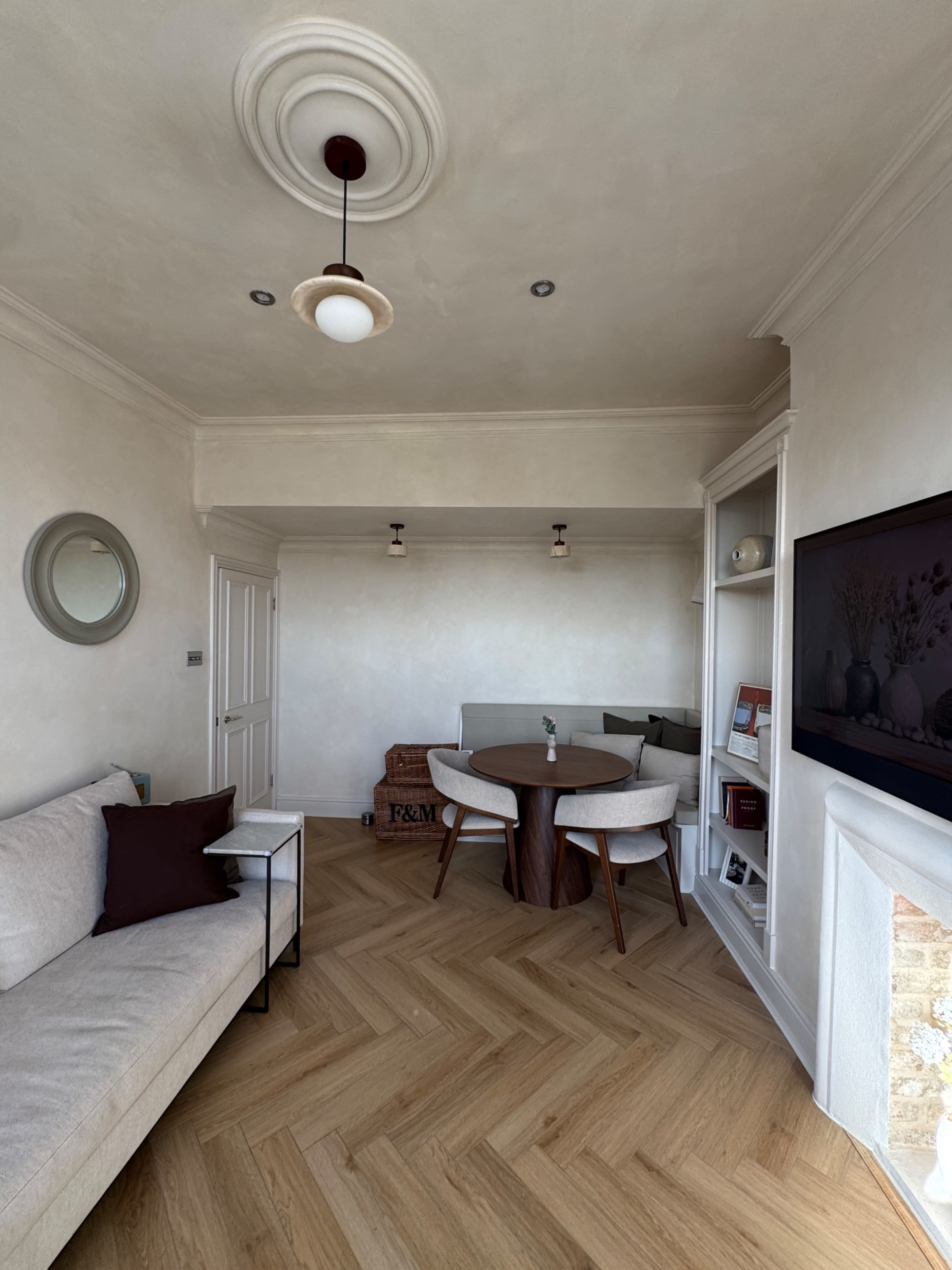 The image shows a light-colored, cozy living space featuring a sofa, a circular dining table with chairs, and built-in shelves.