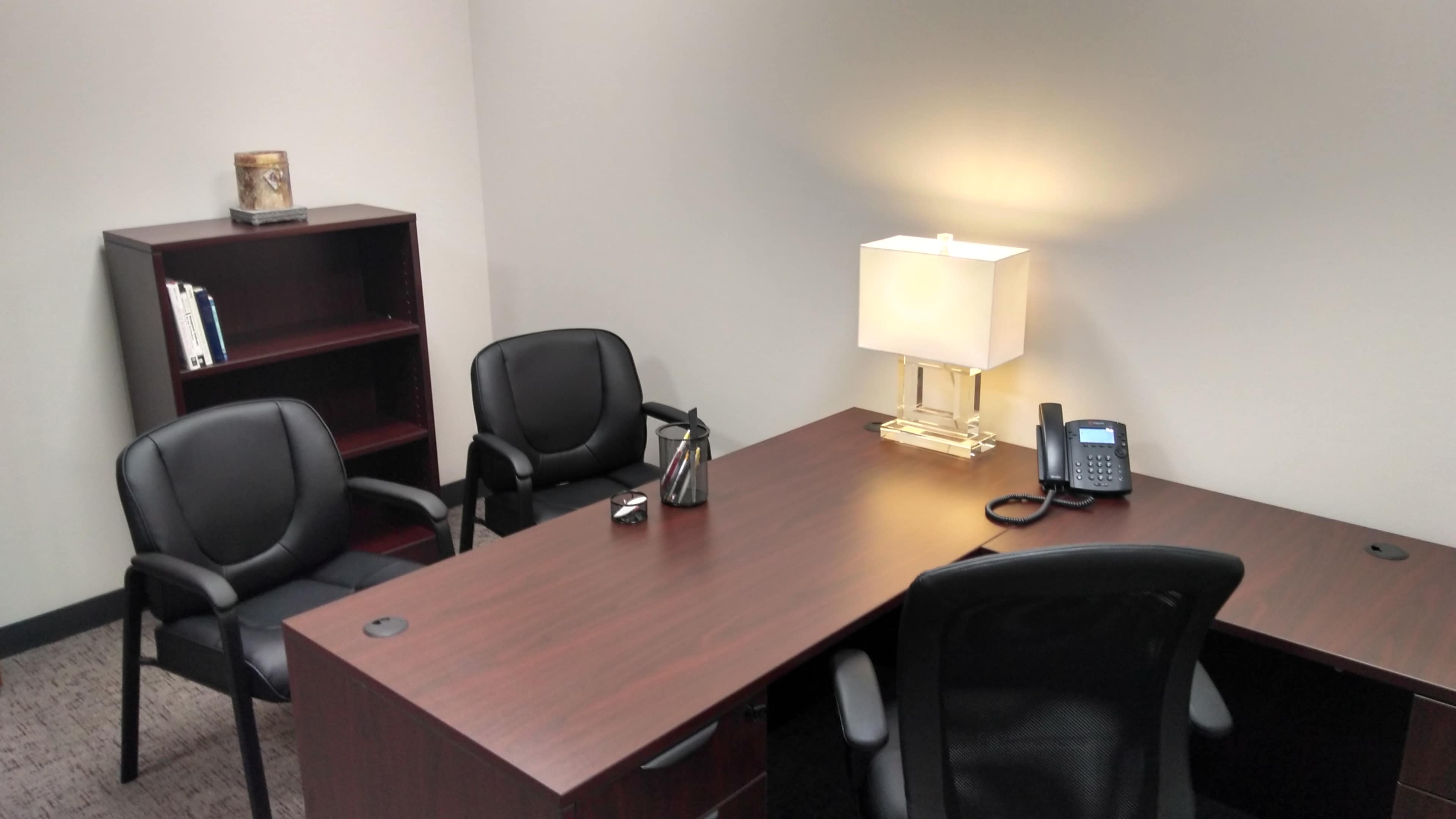The image shows a minimalistic office space with a wooden desk, a lamp, a phone, and two black chairs arranged around a small table.