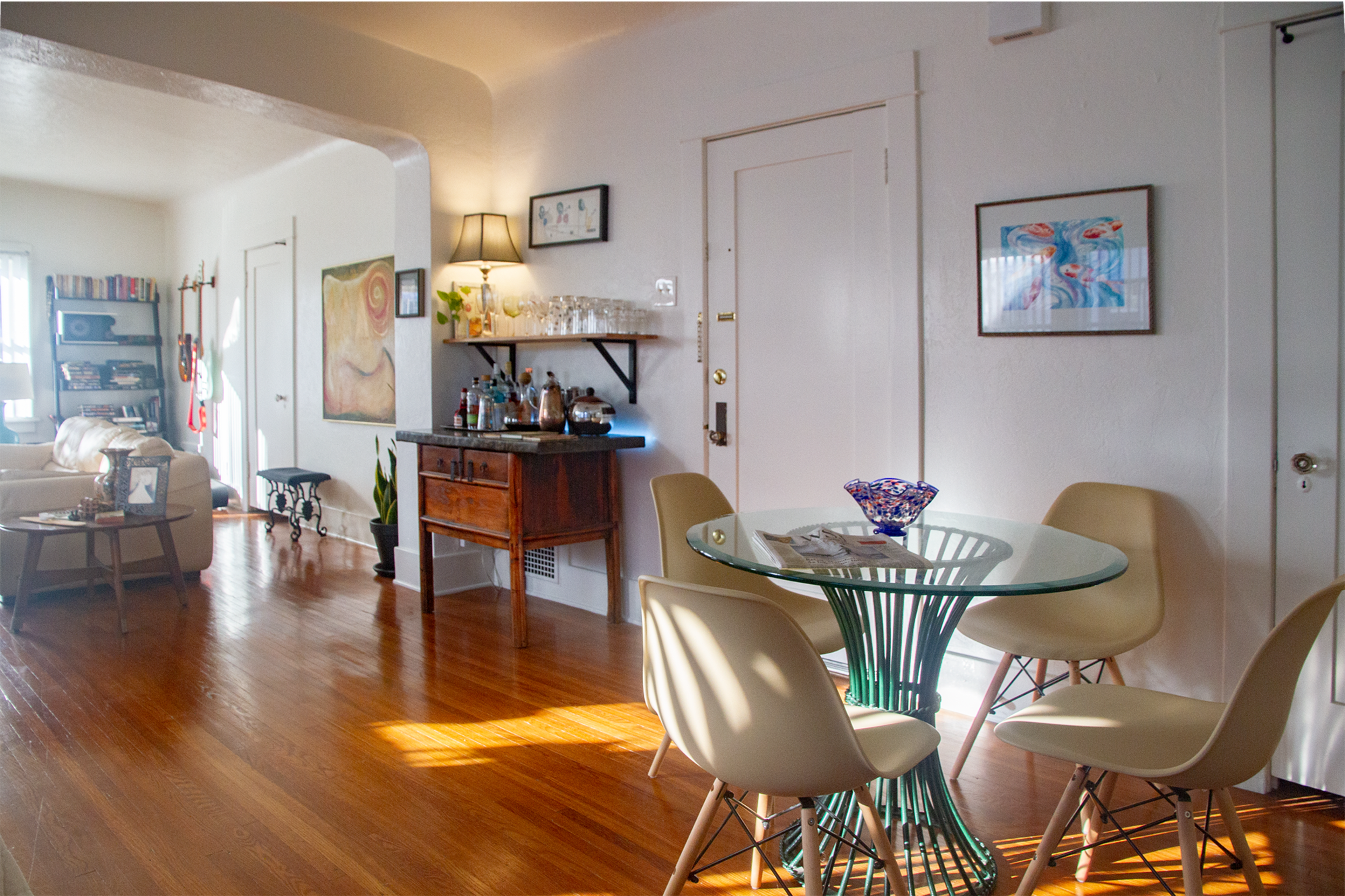 A well-lit interior space featuring a dining table with four chairs, a wooden sideboard, and a cozy seating area with a sofa and shelves filled with books.
