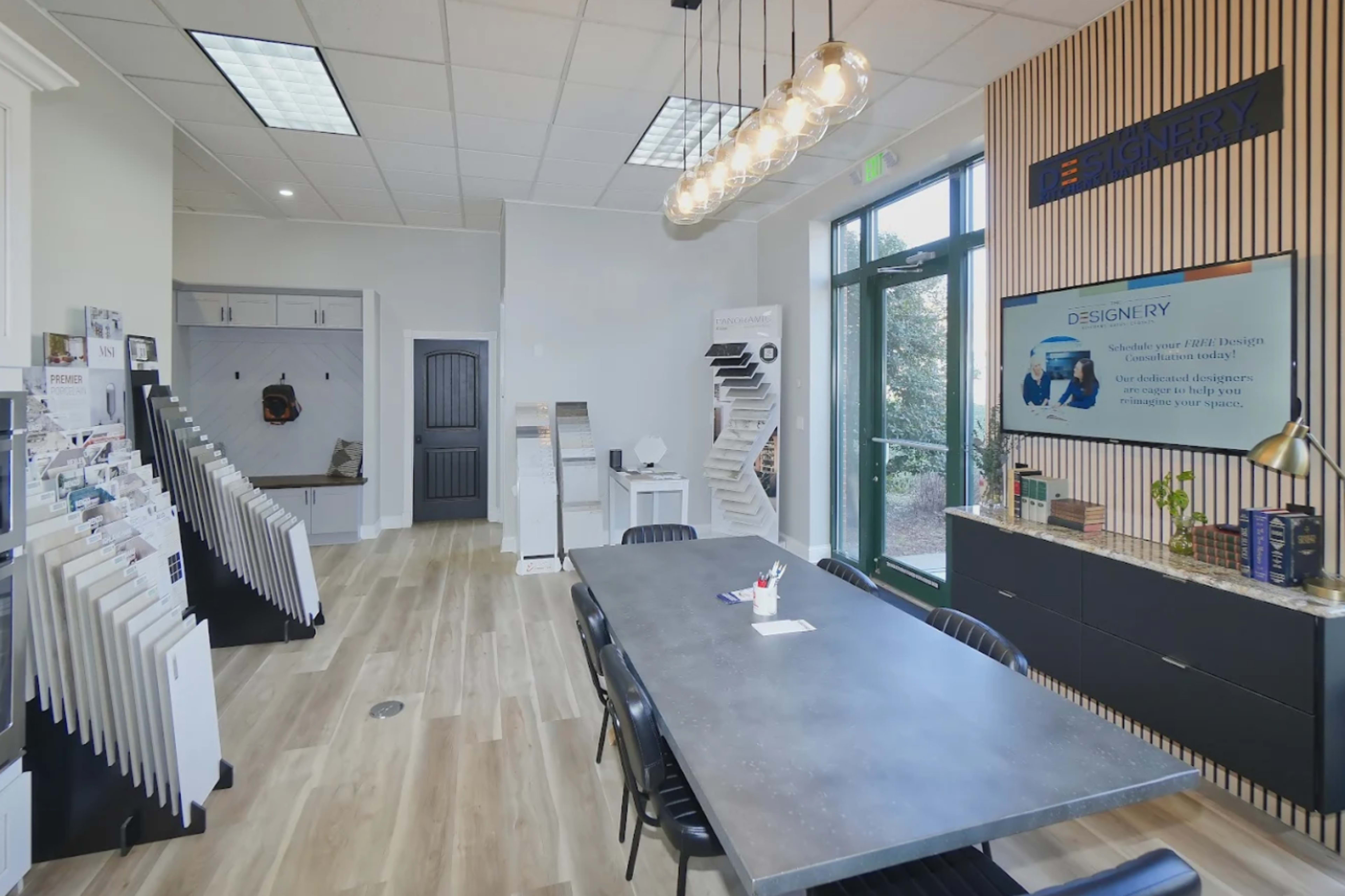 The image shows a modern office space with a large gray table, black chairs, and walls displaying design samples and promotional materials.
