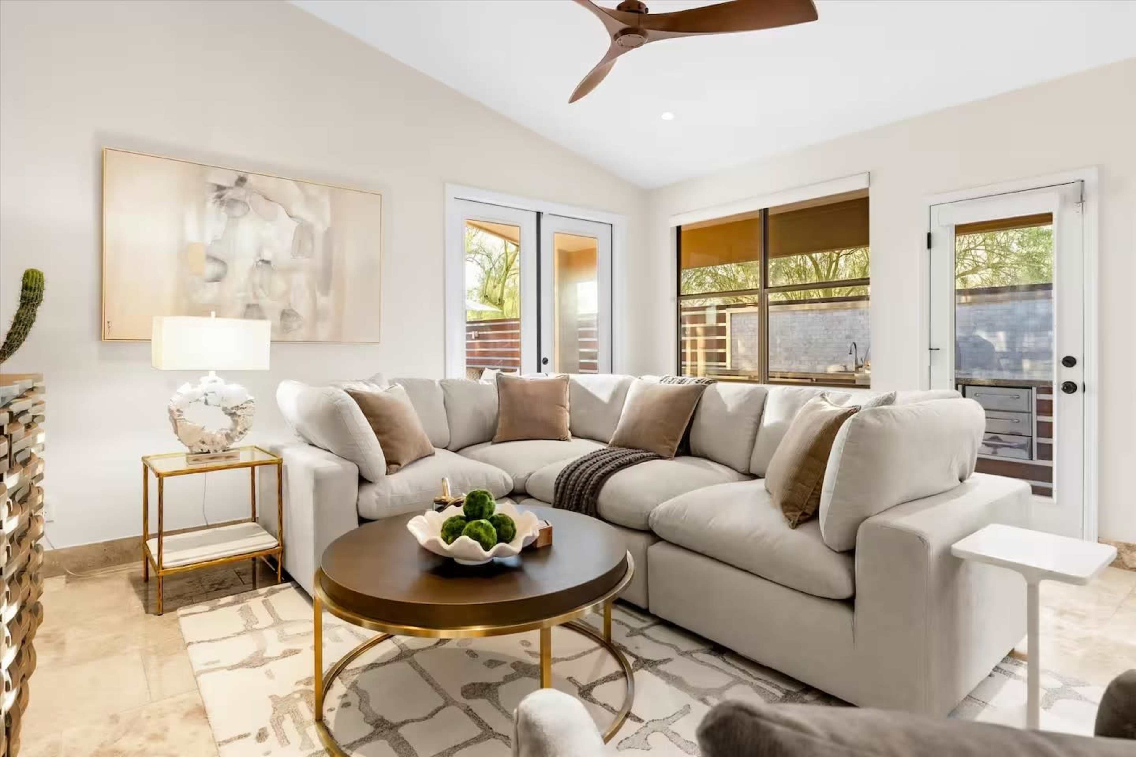 The image shows a cozy living room featuring a large sectional sofa, a round coffee table, and large windows that provide natural light.