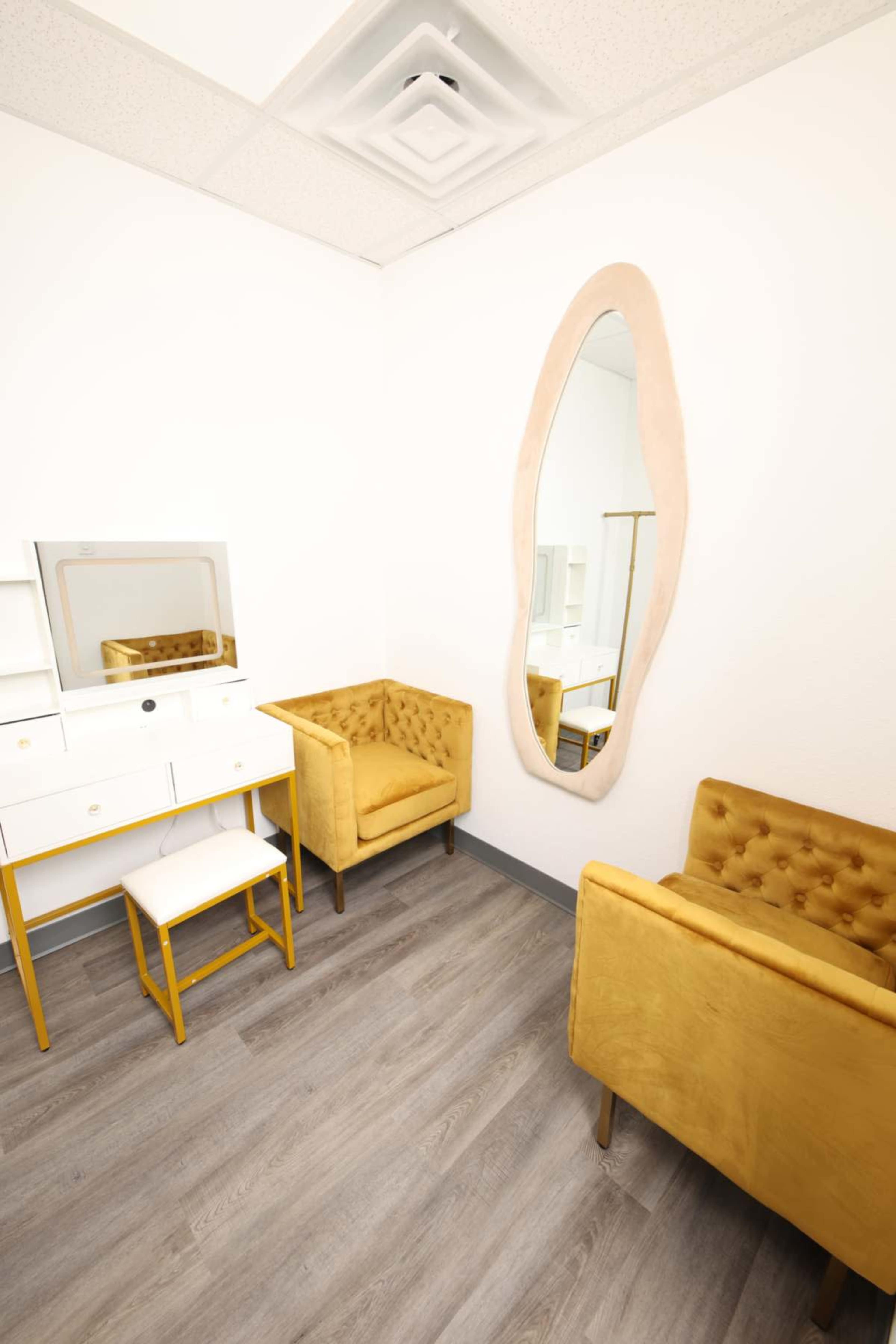 The image shows a small, well-lit room with two yellow armchairs, a white vanity with a mirror, and wooden flooring.