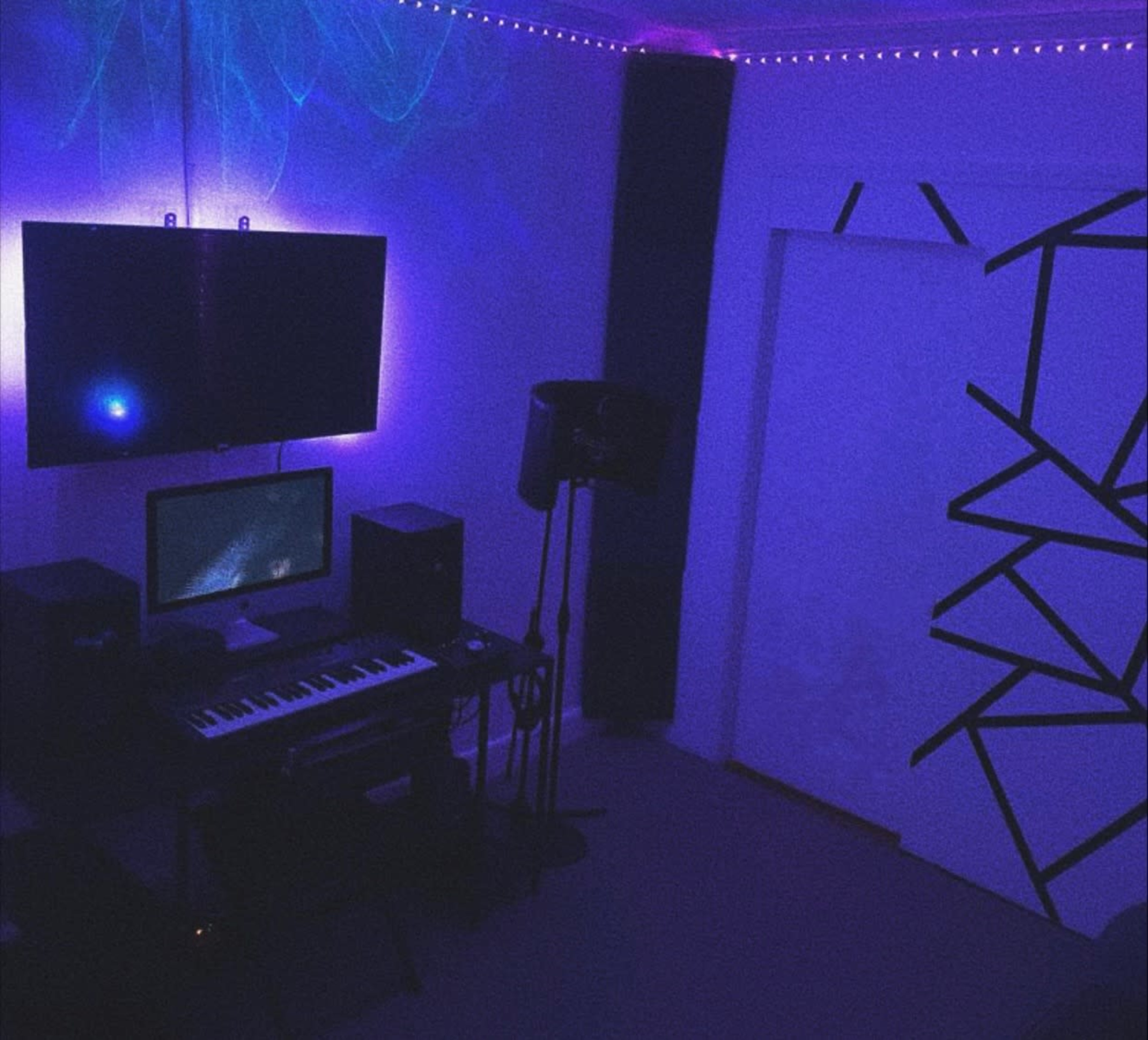 A dimly lit music studio with a keyboard, computer, and monitor set up, surrounded by illuminated walls and a patterned door.