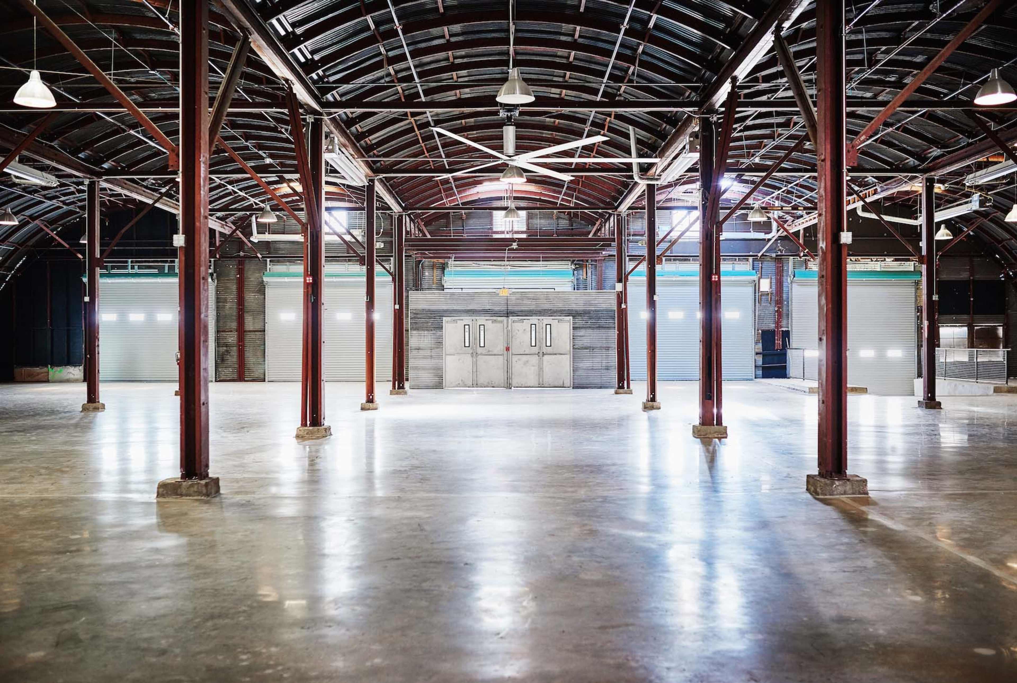 An empty industrial warehouse with large metal pillars, a polished concrete floor, and overhead lights illuminating the space.