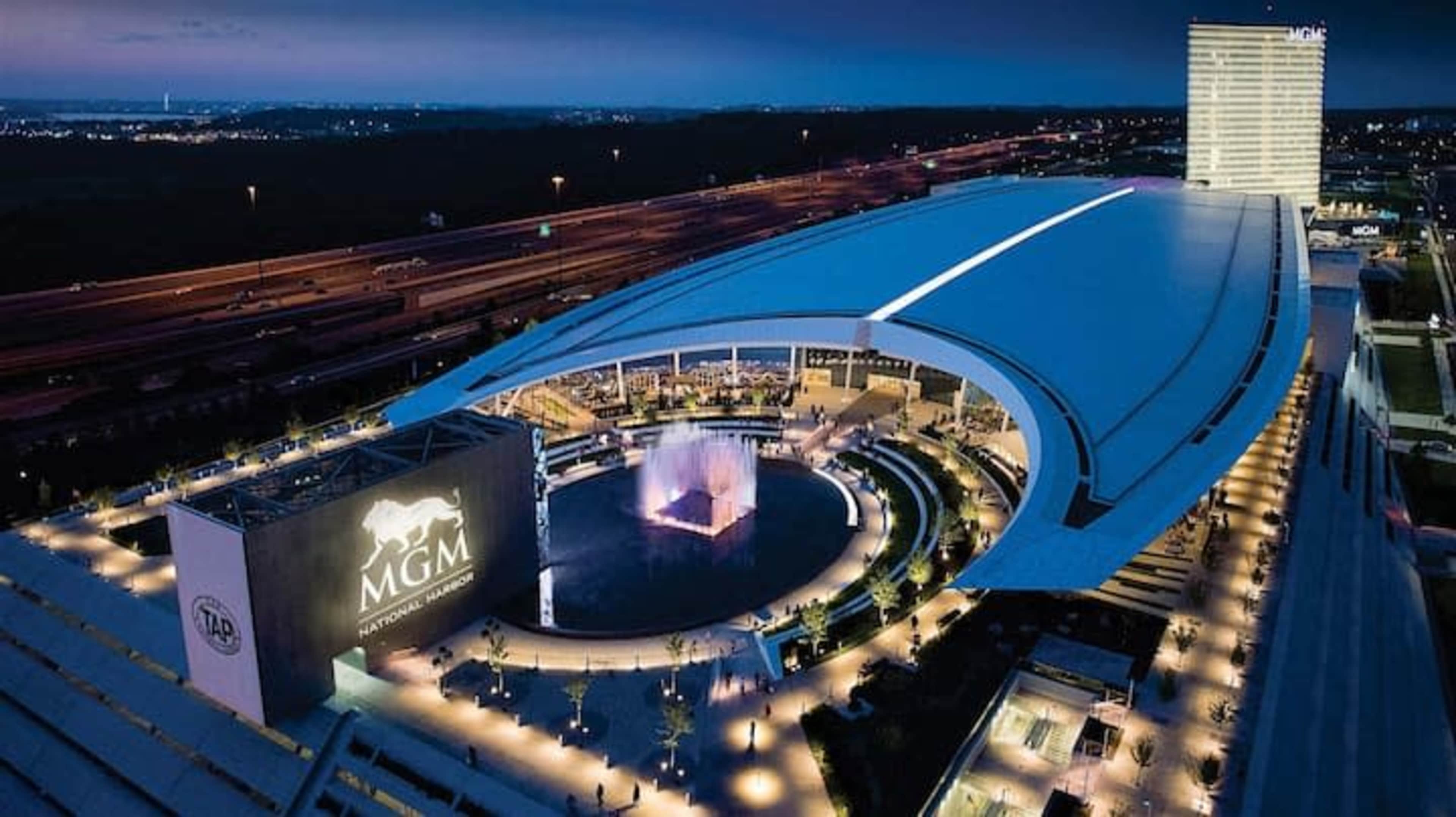 An aerial view of the MGM Grand Casino and Resort at dusk, featuring a large circular fountain in front and a highway in the background.