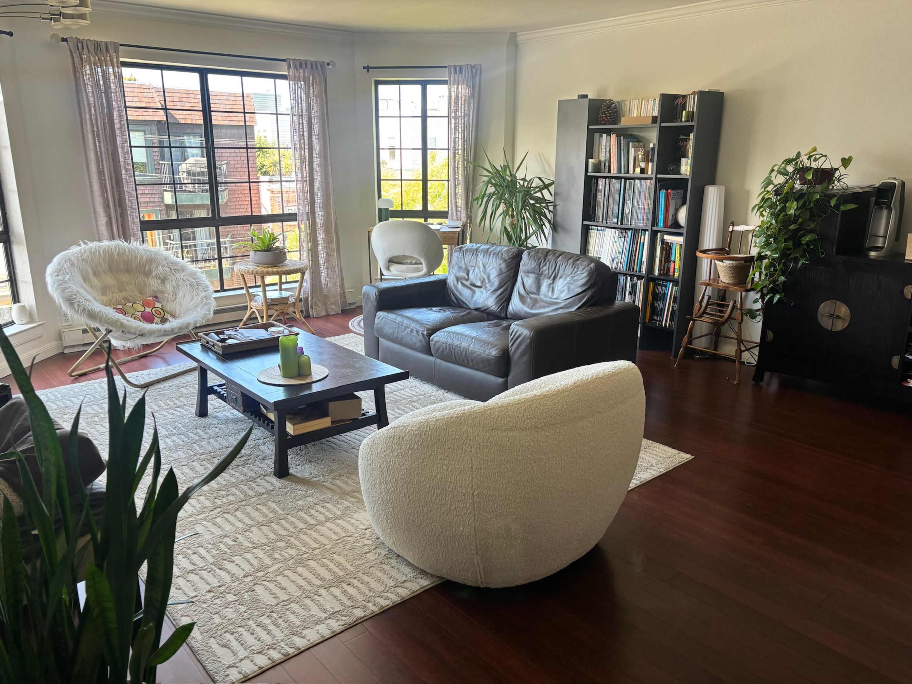 A bright living room features a leather couch, circular chairs, a coffee table, and shelves filled with books, illuminated by large windows.
