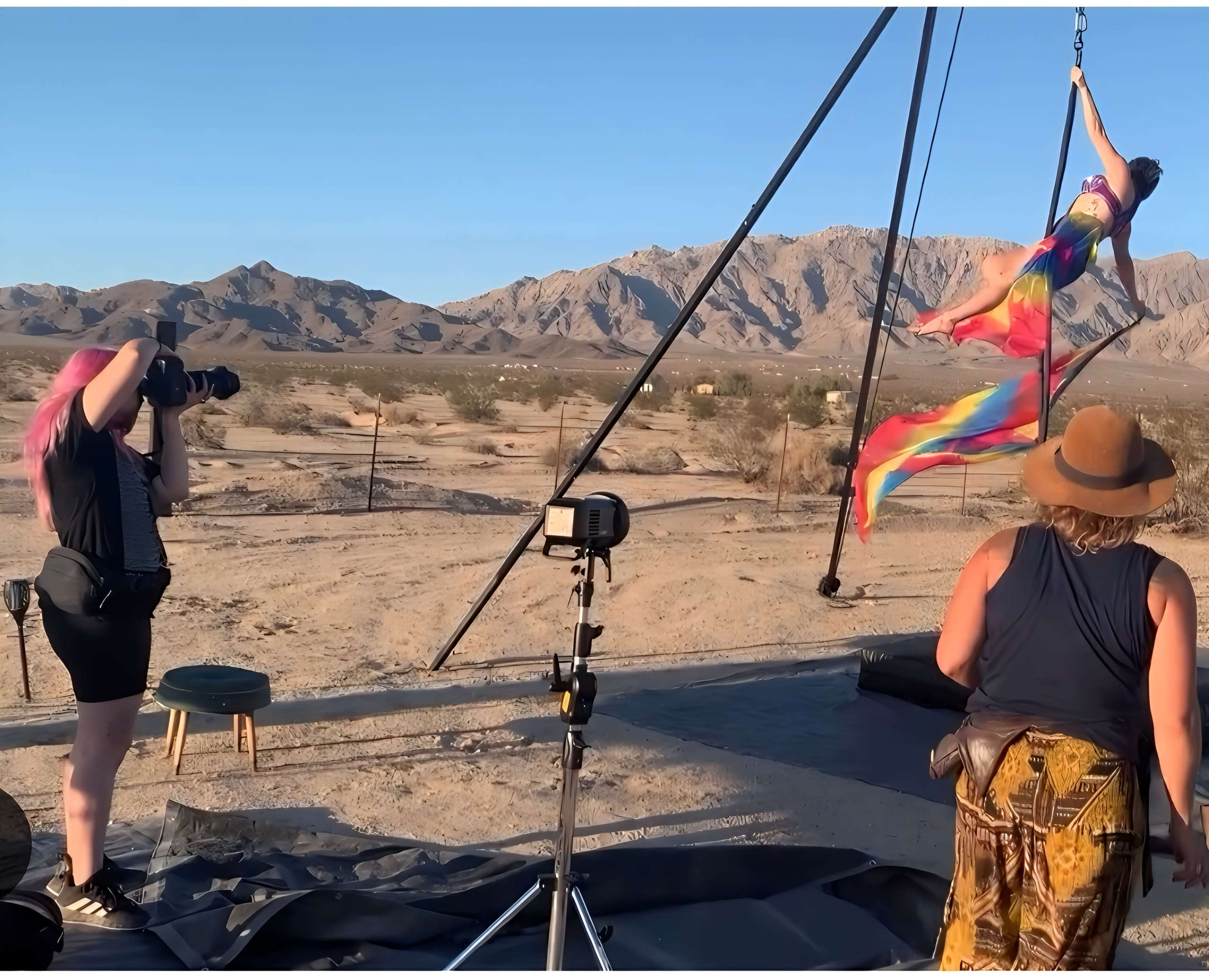 A woman performs aerial acrobatics on a swing in a desert setting while two photographers capture the scene.
