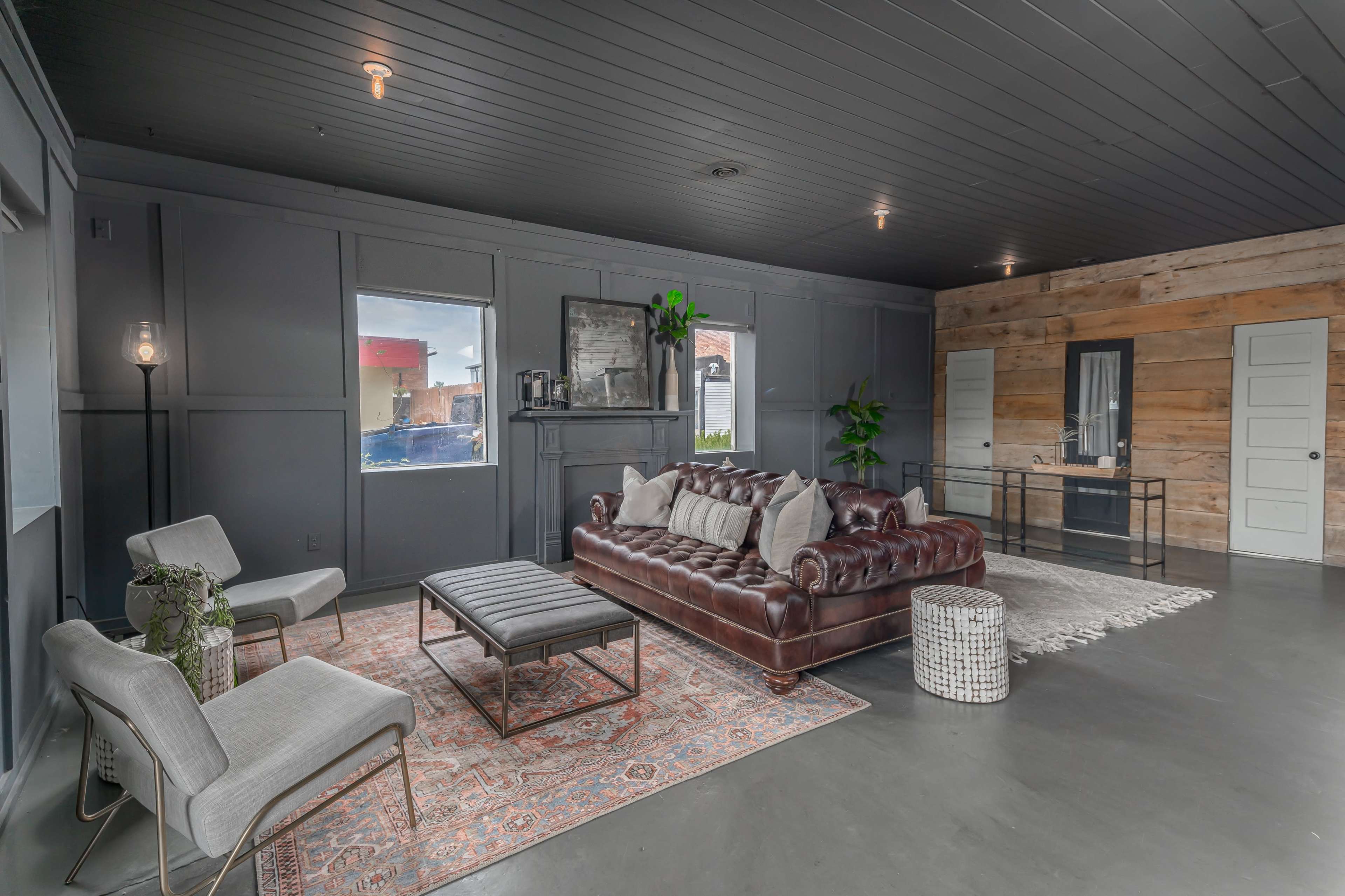 A modern living room with a dark ceiling, a brown leather couch, two light-colored chairs, and a rug on a concrete floor.