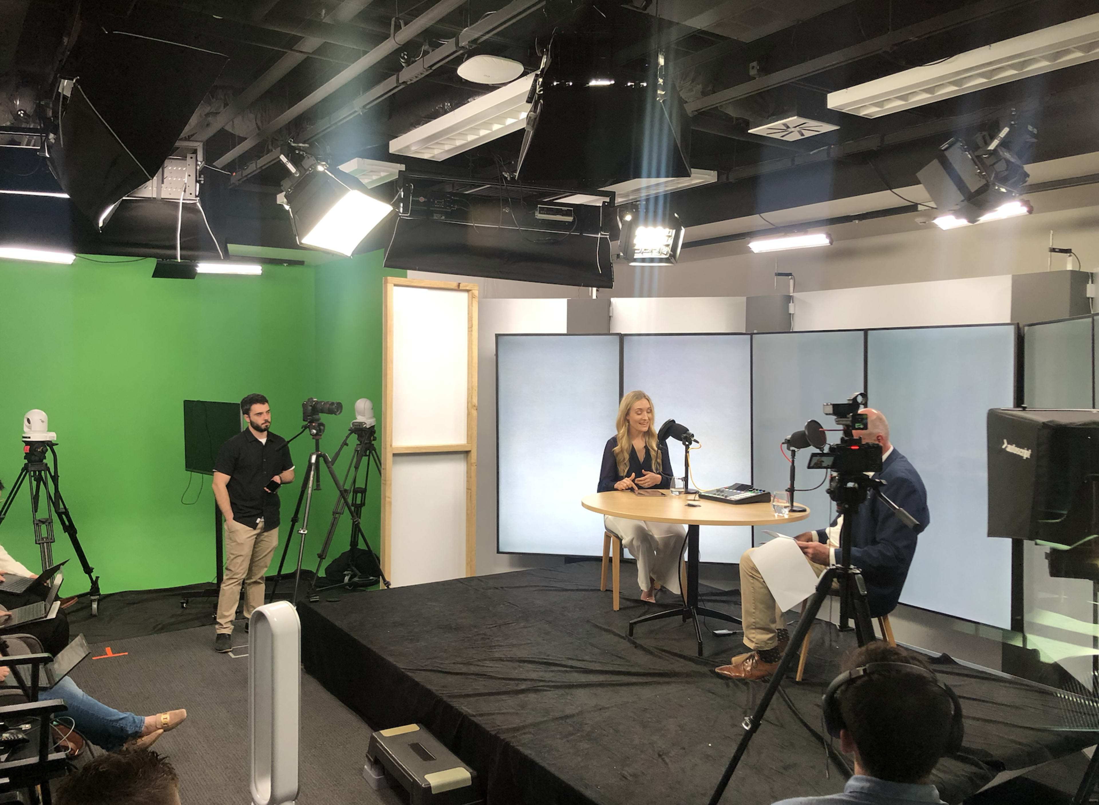 A television studio setting features two people engaged in an interview, with multiple cameras, lighting equipment, and a green screen in the background.