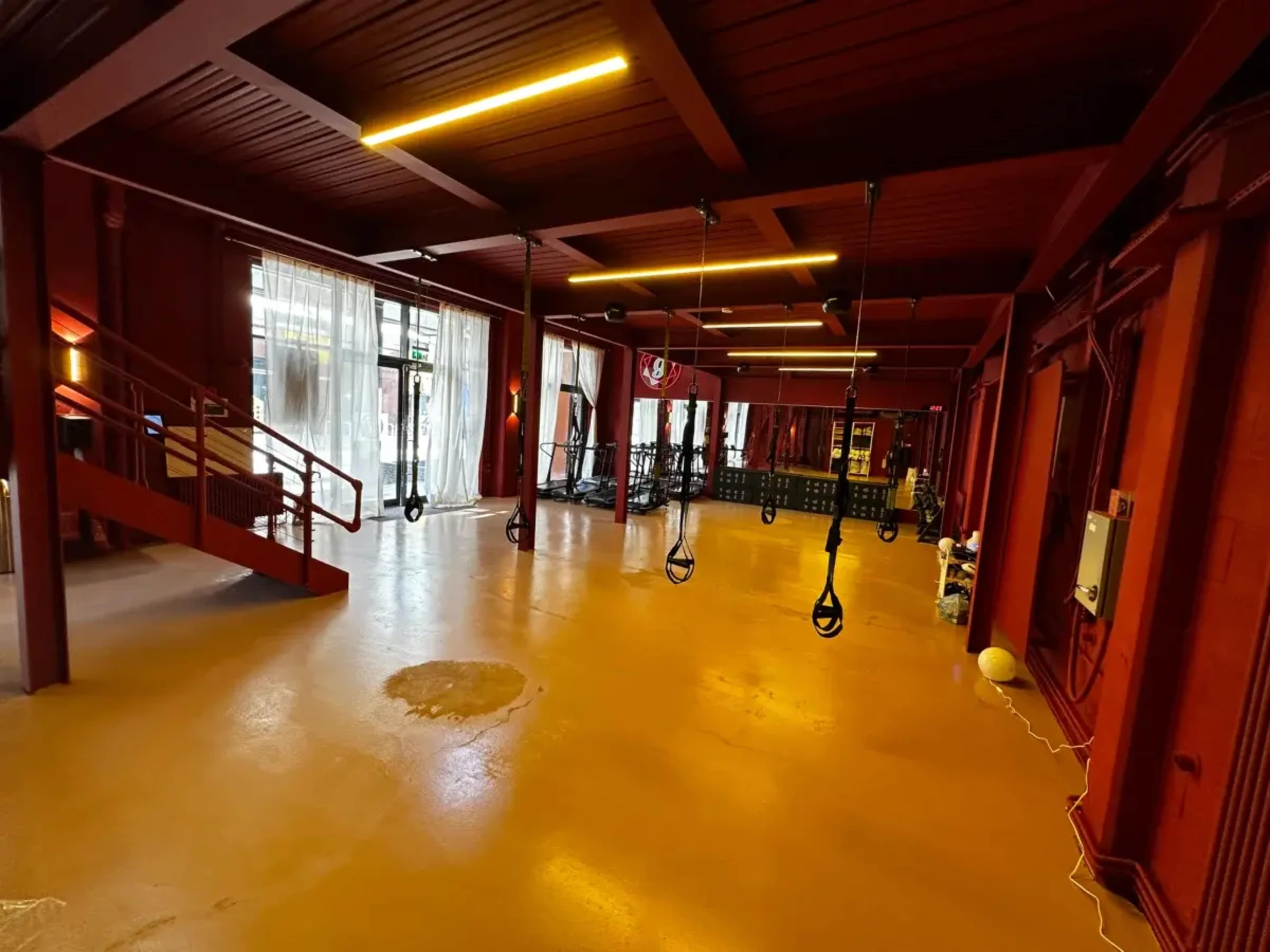 The image shows a spacious indoor gym with wooden walls, bright overhead lights, and various fitness equipment arranged along one side.