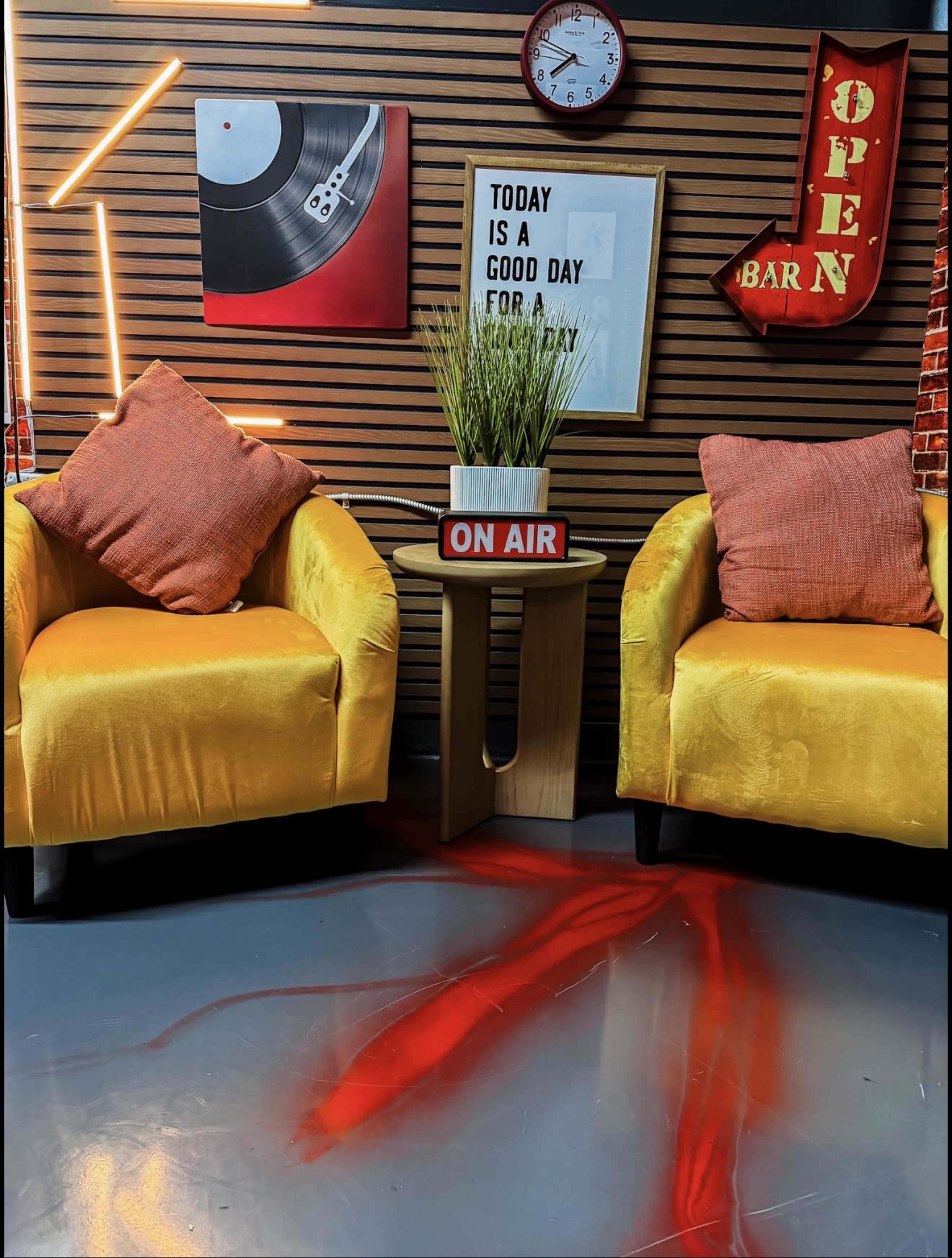 The scene features two yellow armchairs with decorative pillows, a small table displaying an "ON AIR" sign, and a backdrop of wall art and neon lighting.