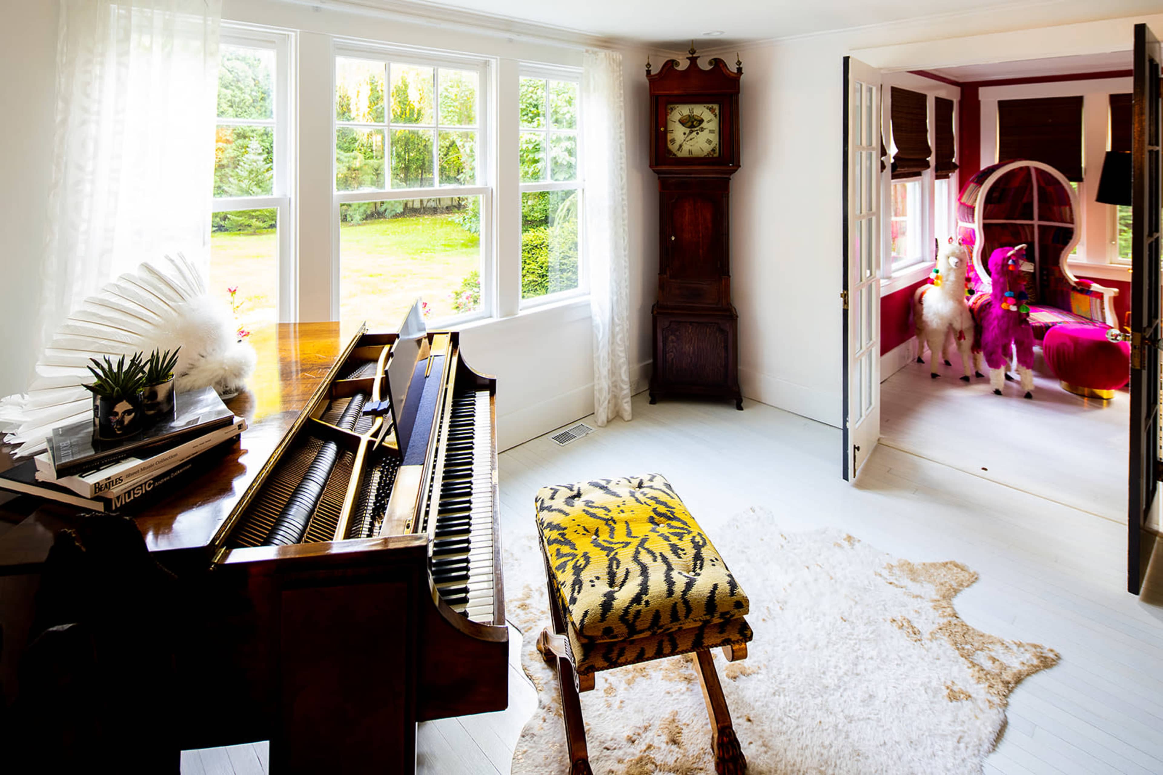 A grand piano and an ornate grandfather clock are situated in a bright room that opens into another space featuring colorful seating and two decorative unicorns.