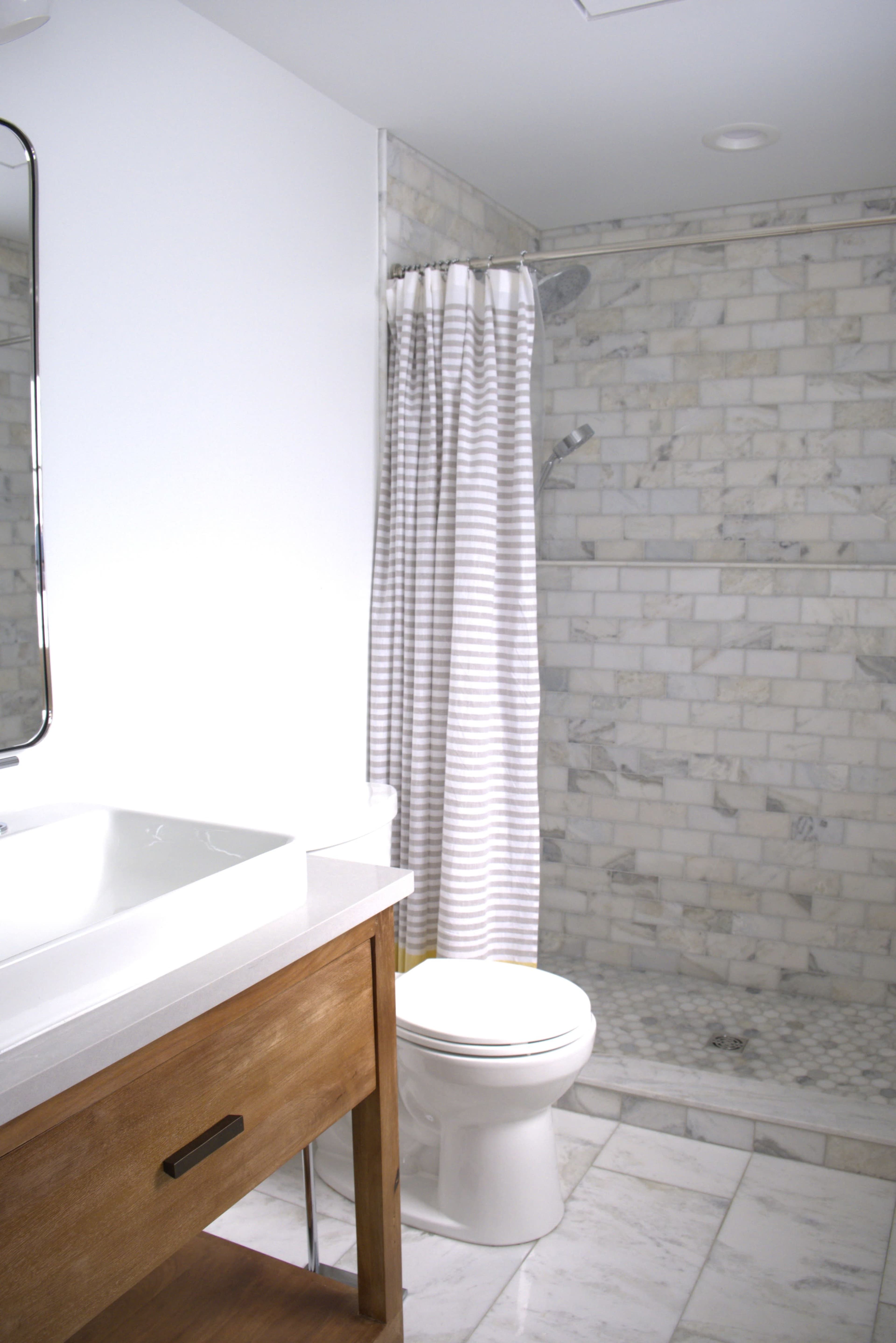 A modern bathroom features a shower with a striped curtain, a white toilet, and a wooden vanity with a sink and a large mirror.