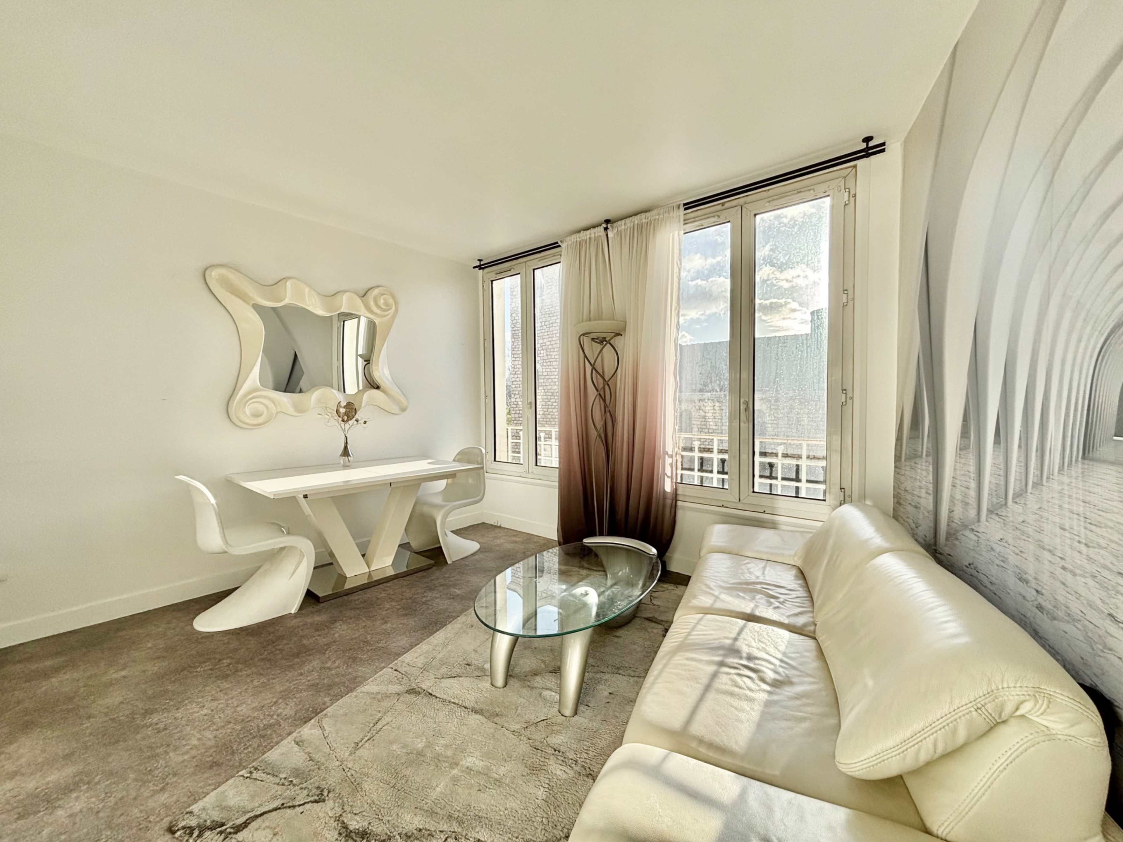 The image shows a modern living room with a white sofa, a glass coffee table, and a dining area featuring a sleek table and chairs beside large windows.