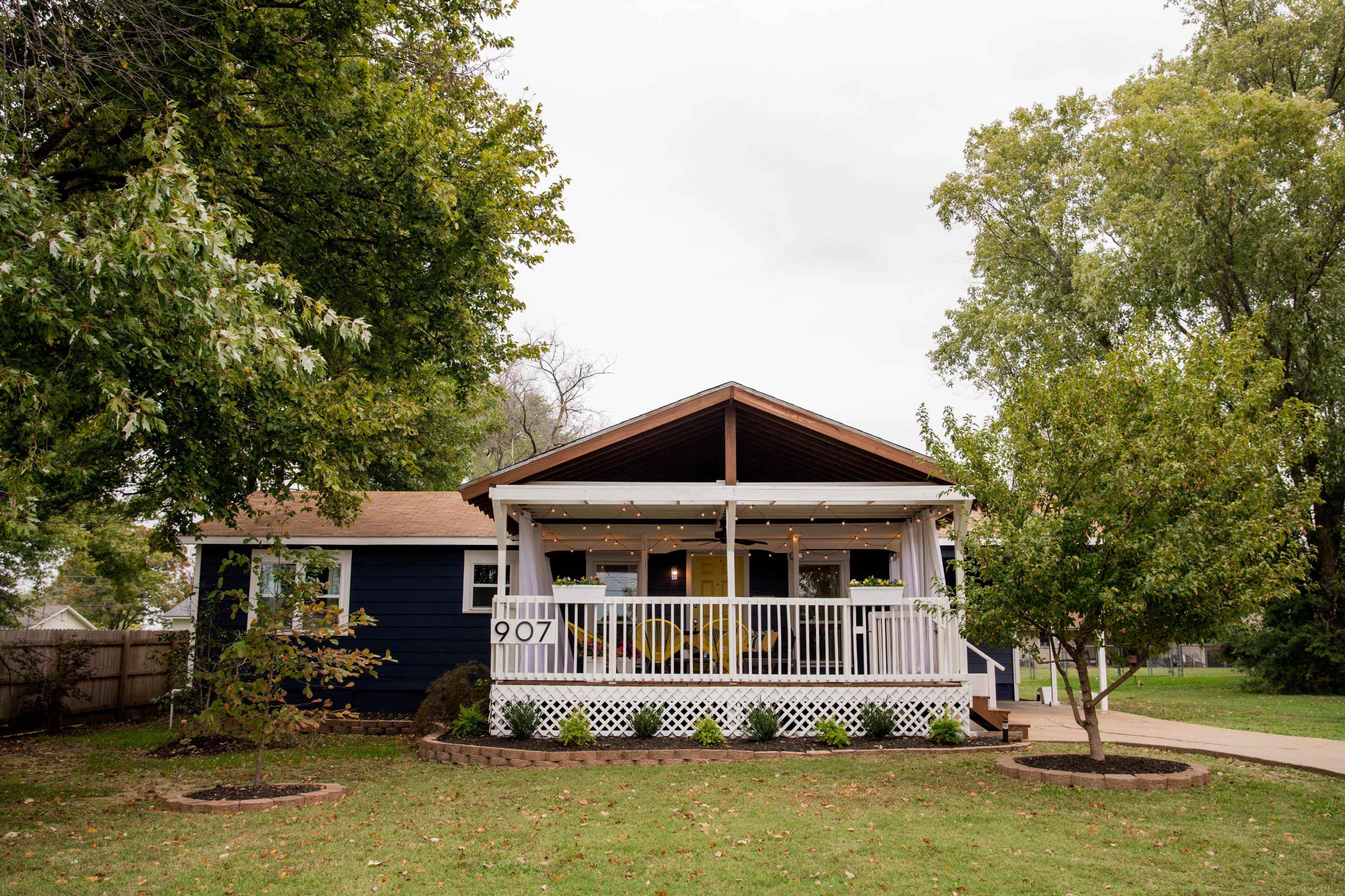A blue house with a white porch, string lights, and a green lawn is situated at 907 on a cloudy day.