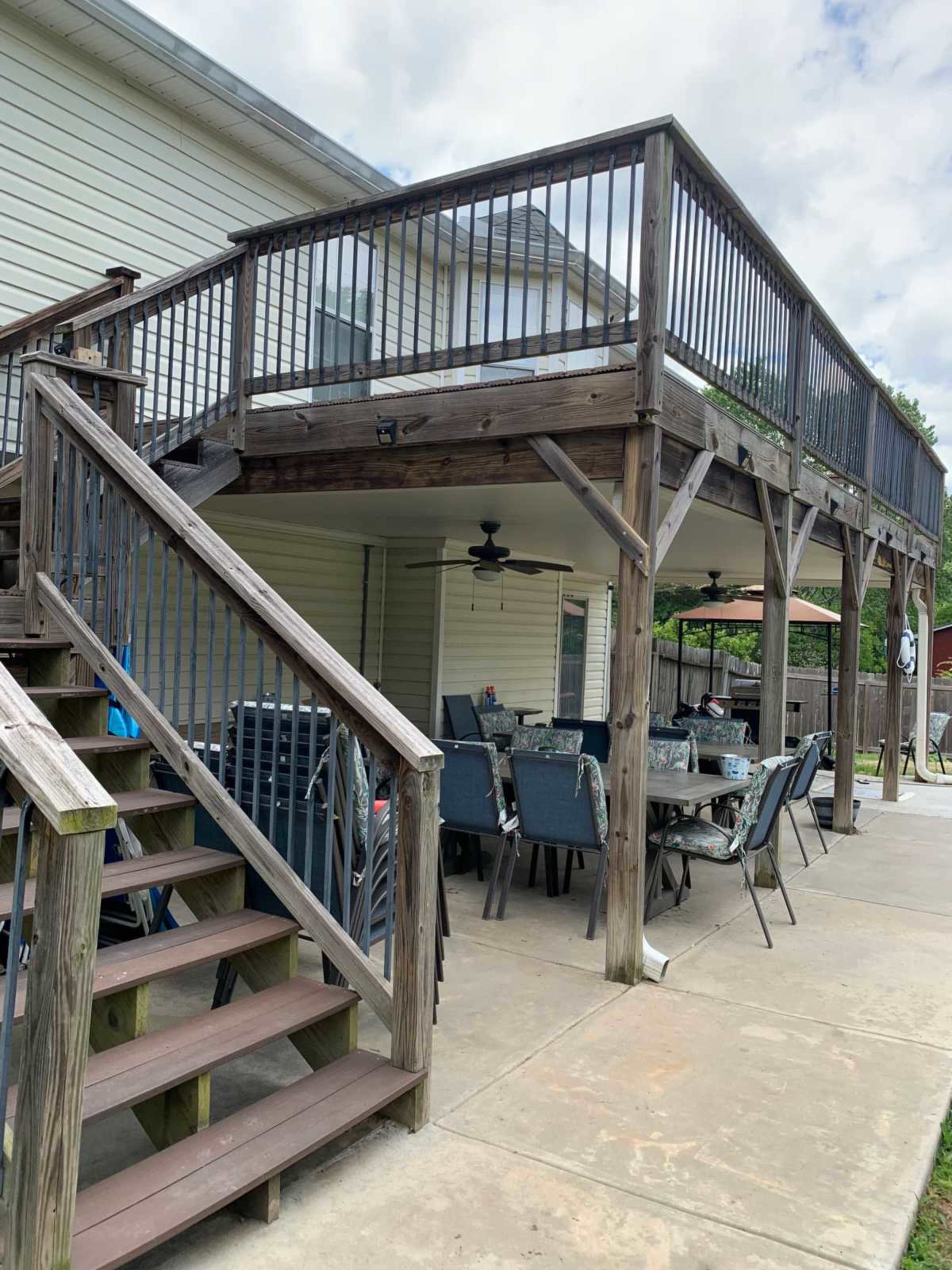 The image shows a wooden deck with a dining area underneath, featuring a table and chairs, adjacent to a house and stairs leading to the upper level.