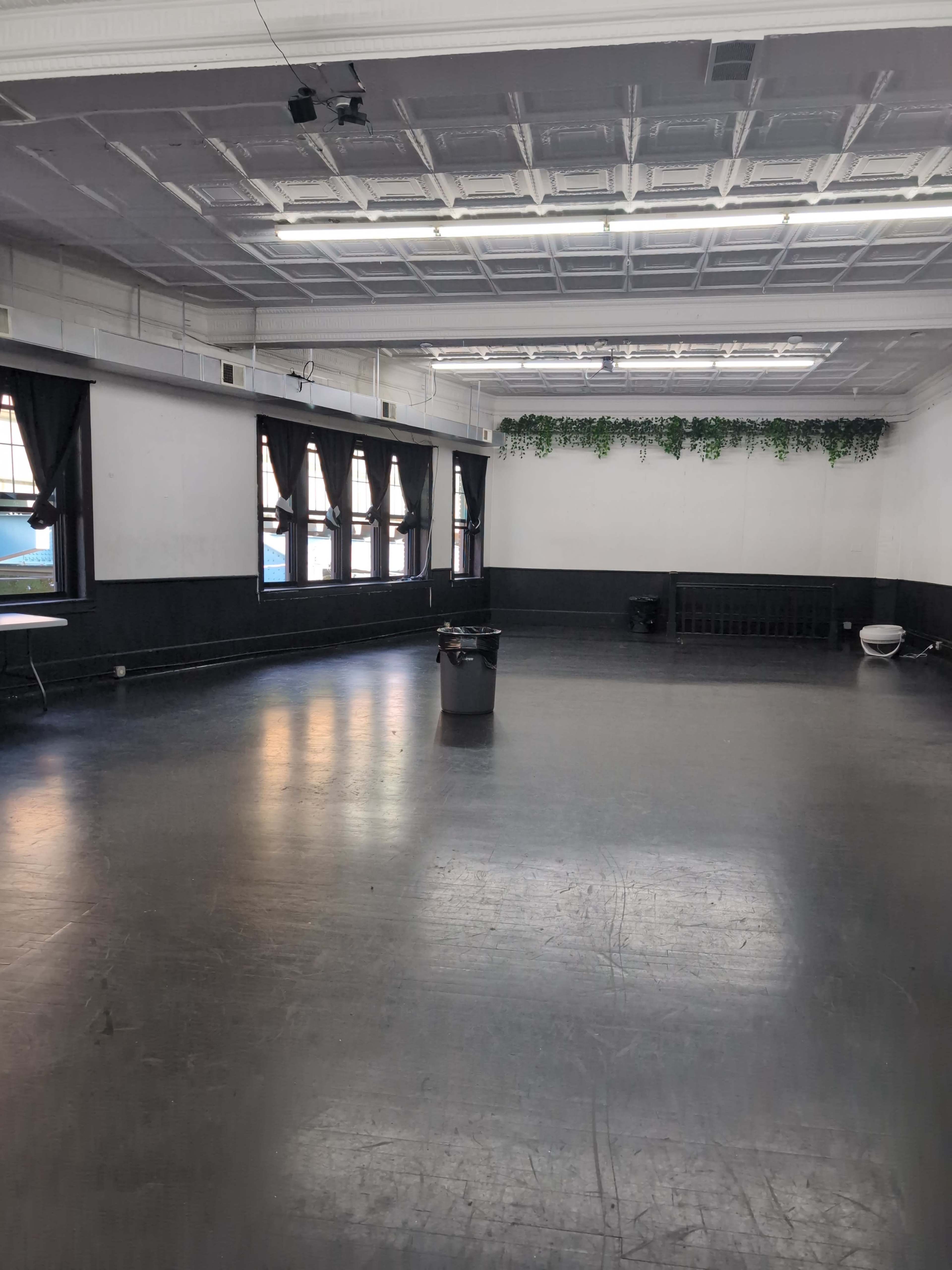 The image shows a spacious, empty dance studio with large windows and black walls, featuring a trash can in the center of the floor.