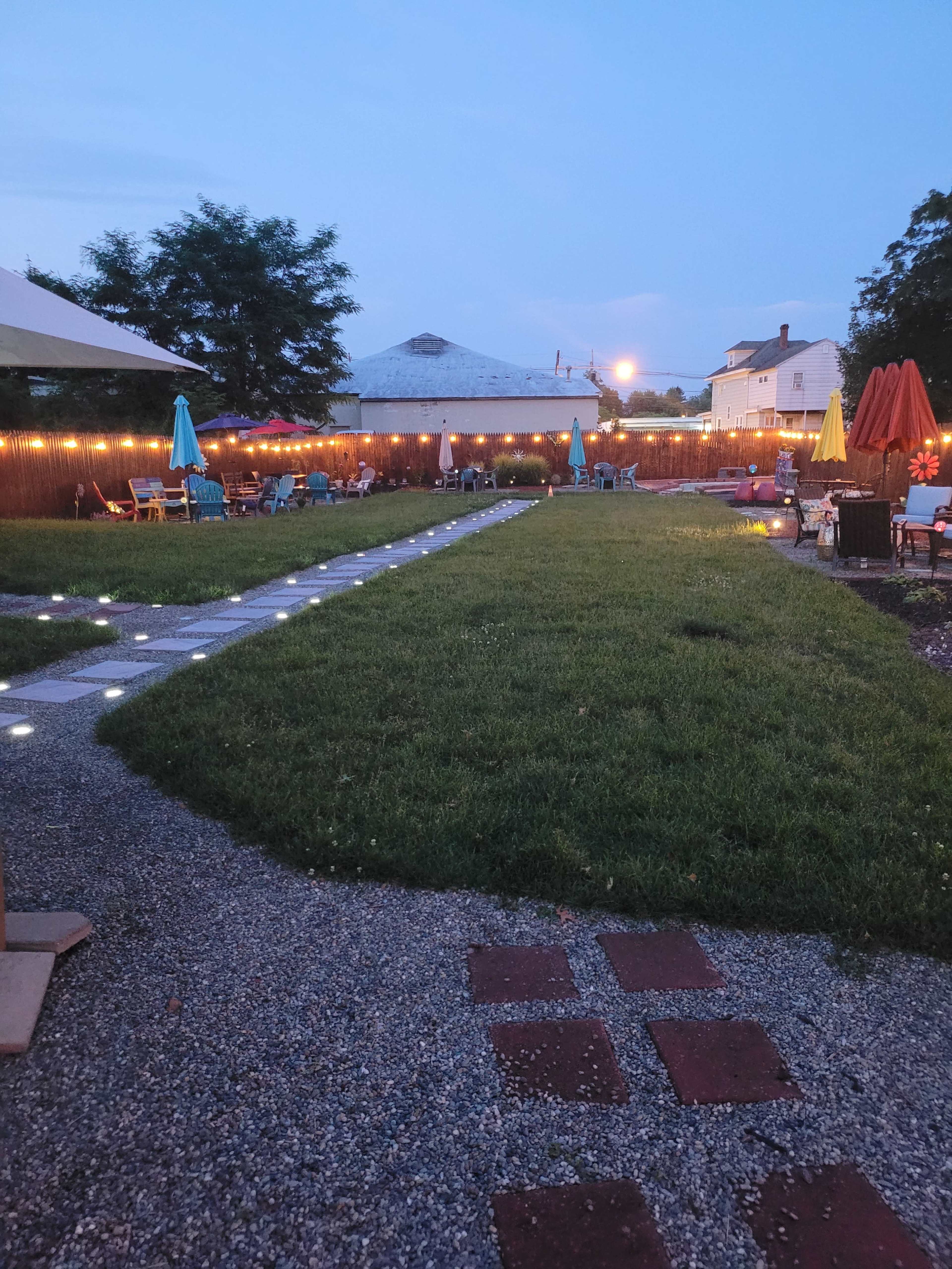 The image shows a well-lit outdoor area with a grassy space, garden furniture, and colorful umbrellas arranged along a pathway.