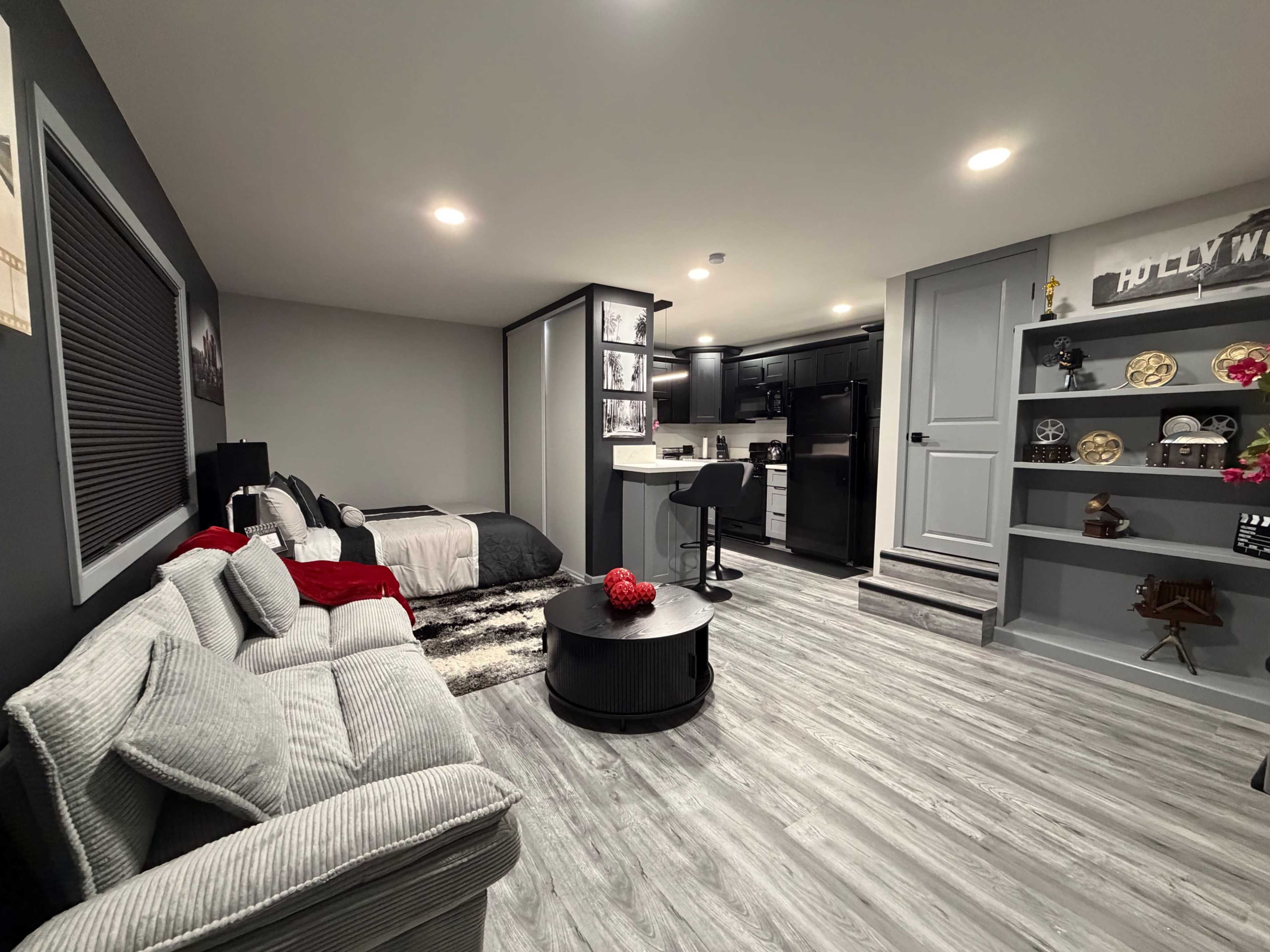 The image shows a modern, minimalistic basement living space with a bed, a cozy couch, and a kitchenette area, accented by decorative shelves.
