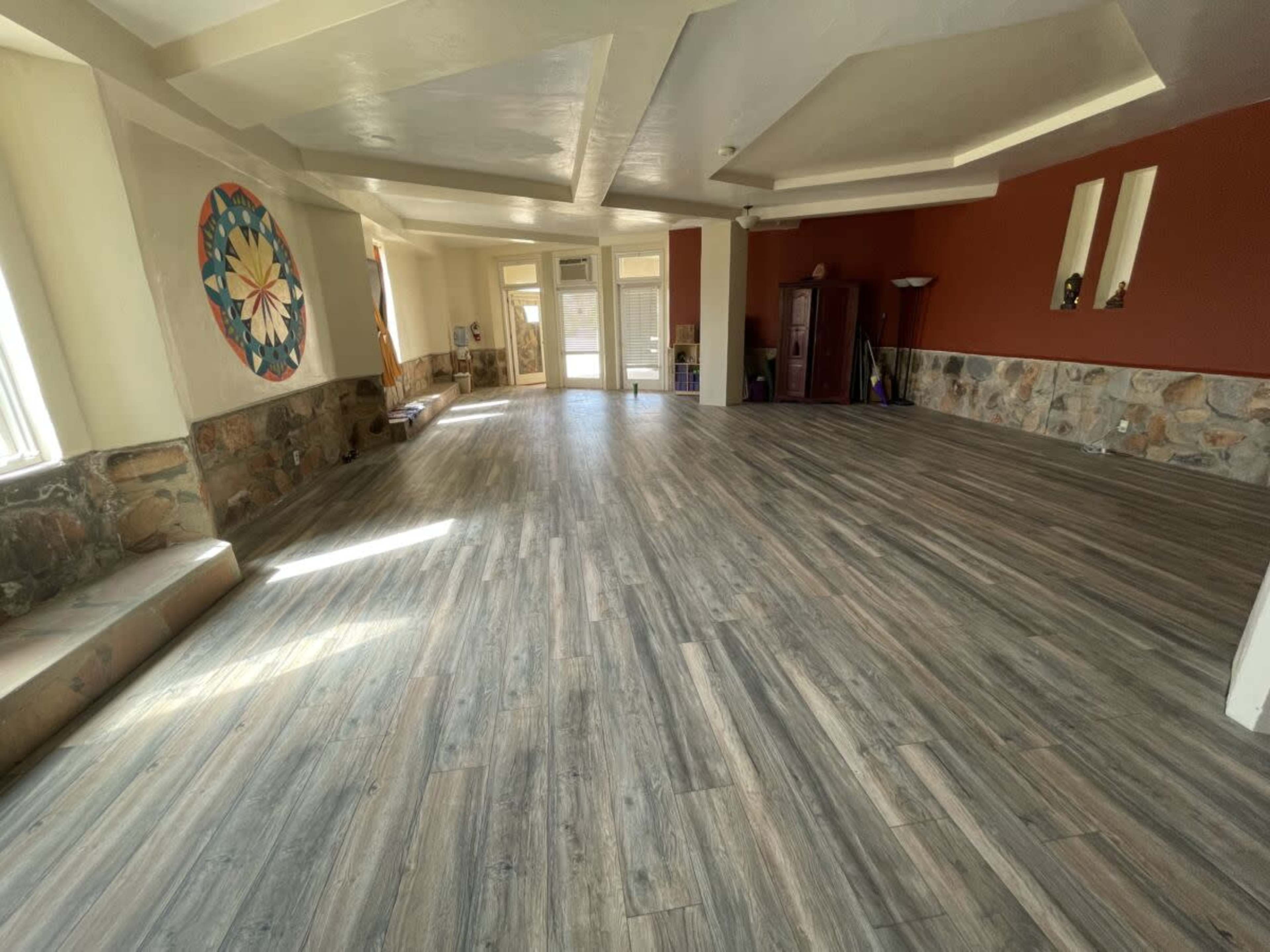 The image shows a spacious, empty room with wooden flooring, stone walls on one side, and large windows letting in natural light.