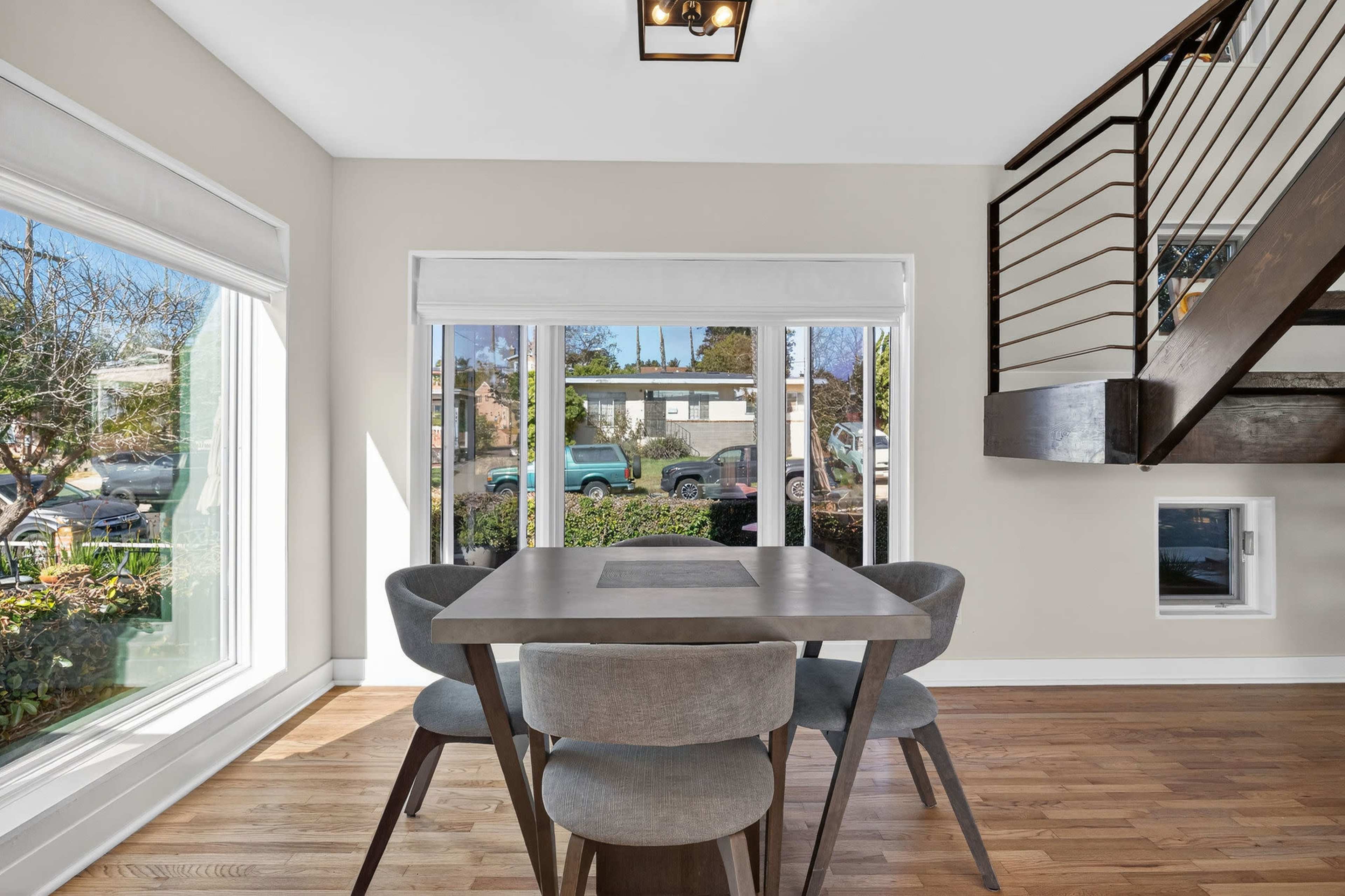 A dining area features a table with four chairs, large windows overlooking a street, and a staircase to the right.