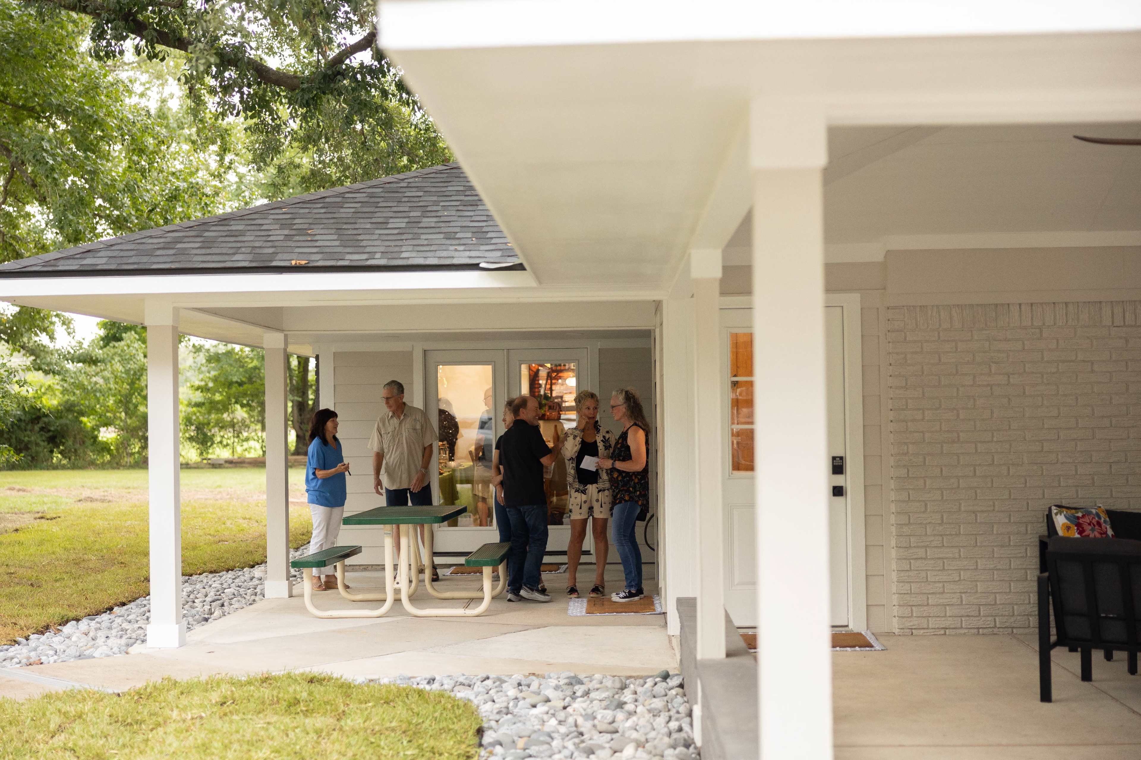 A group of five people is gathered outside a house with a porch, chatting near a picnic table.