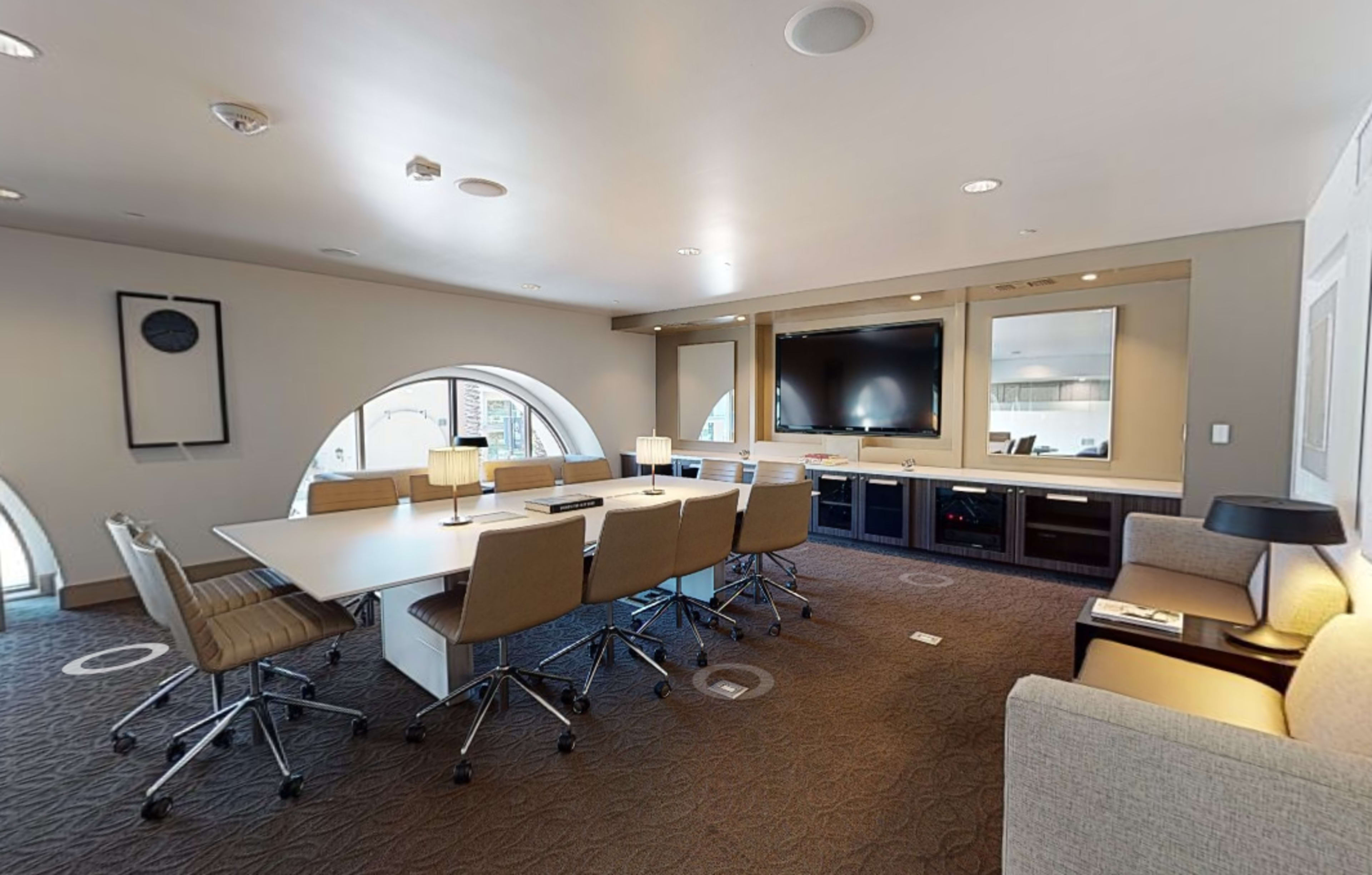 A modern conference room features a large table surrounded by ergonomic chairs, with windows offering natural light and a wall-mounted TV.