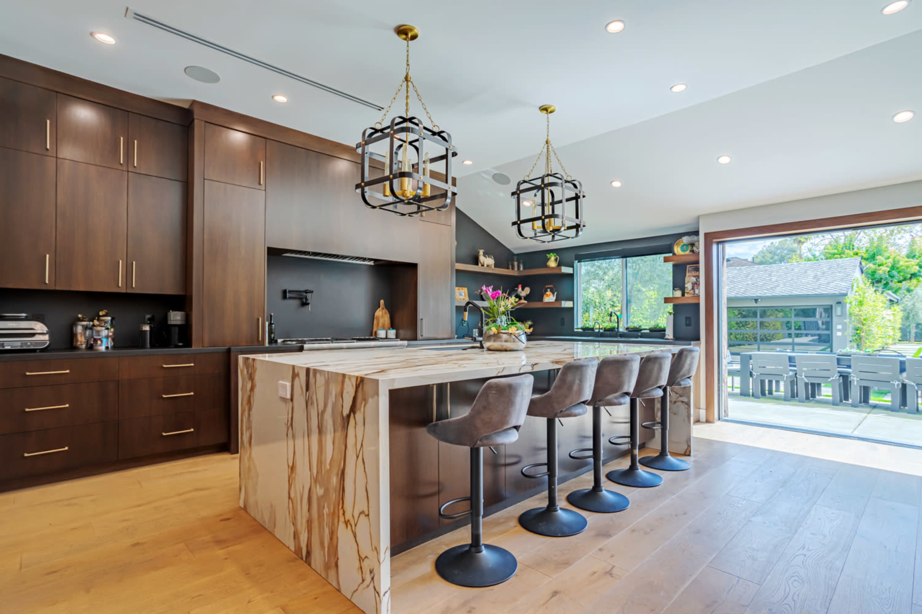 The image shows a modern kitchen with a large marble island, wooden cabinetry, and seating for four at a breakfast bar, adjacent to a glass door leading to an outdoor patio.