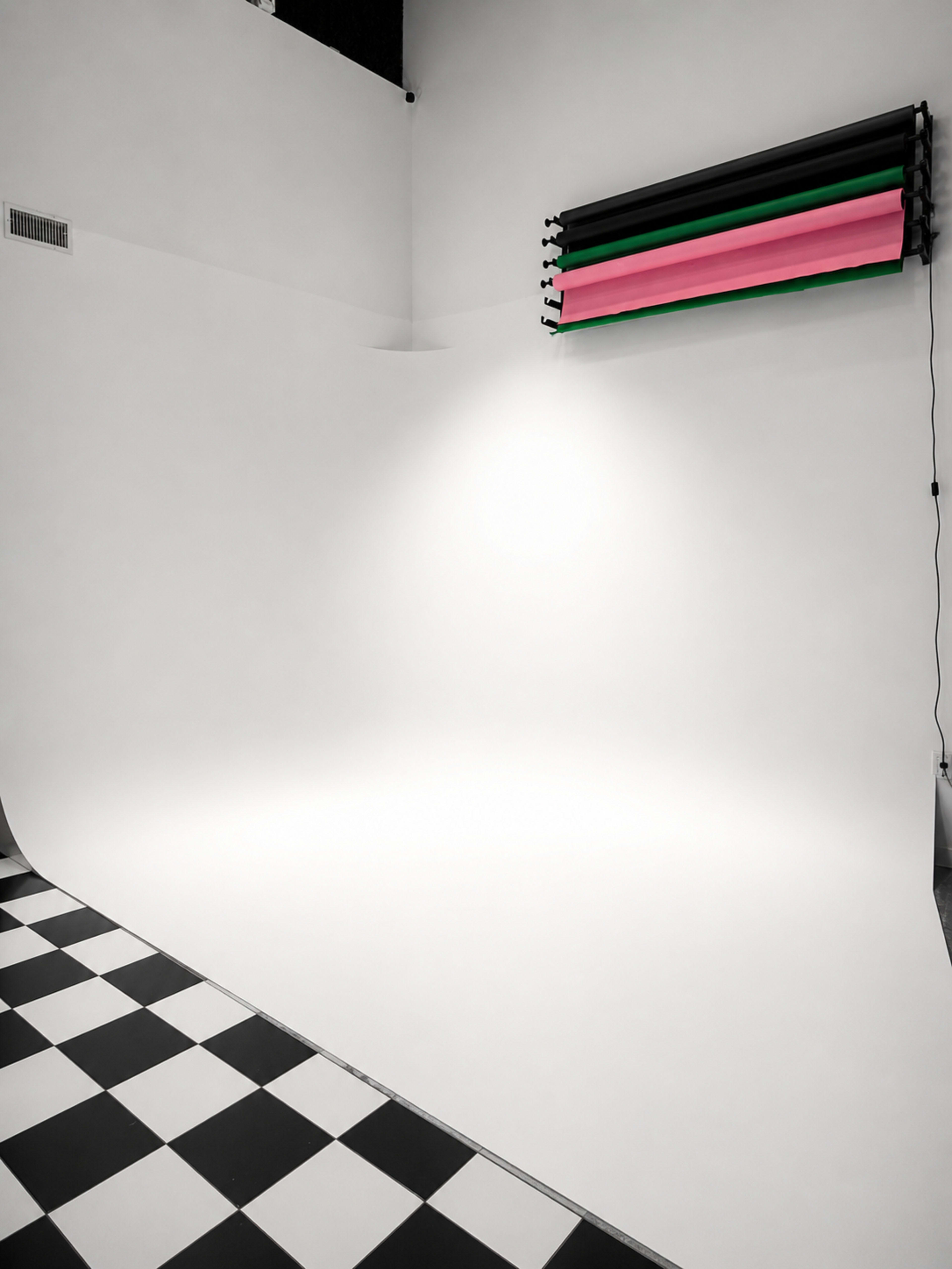 The image shows a studio space with a white backdrop and a black and white checked floor, featuring colored backdrop rolls mounted on the wall.