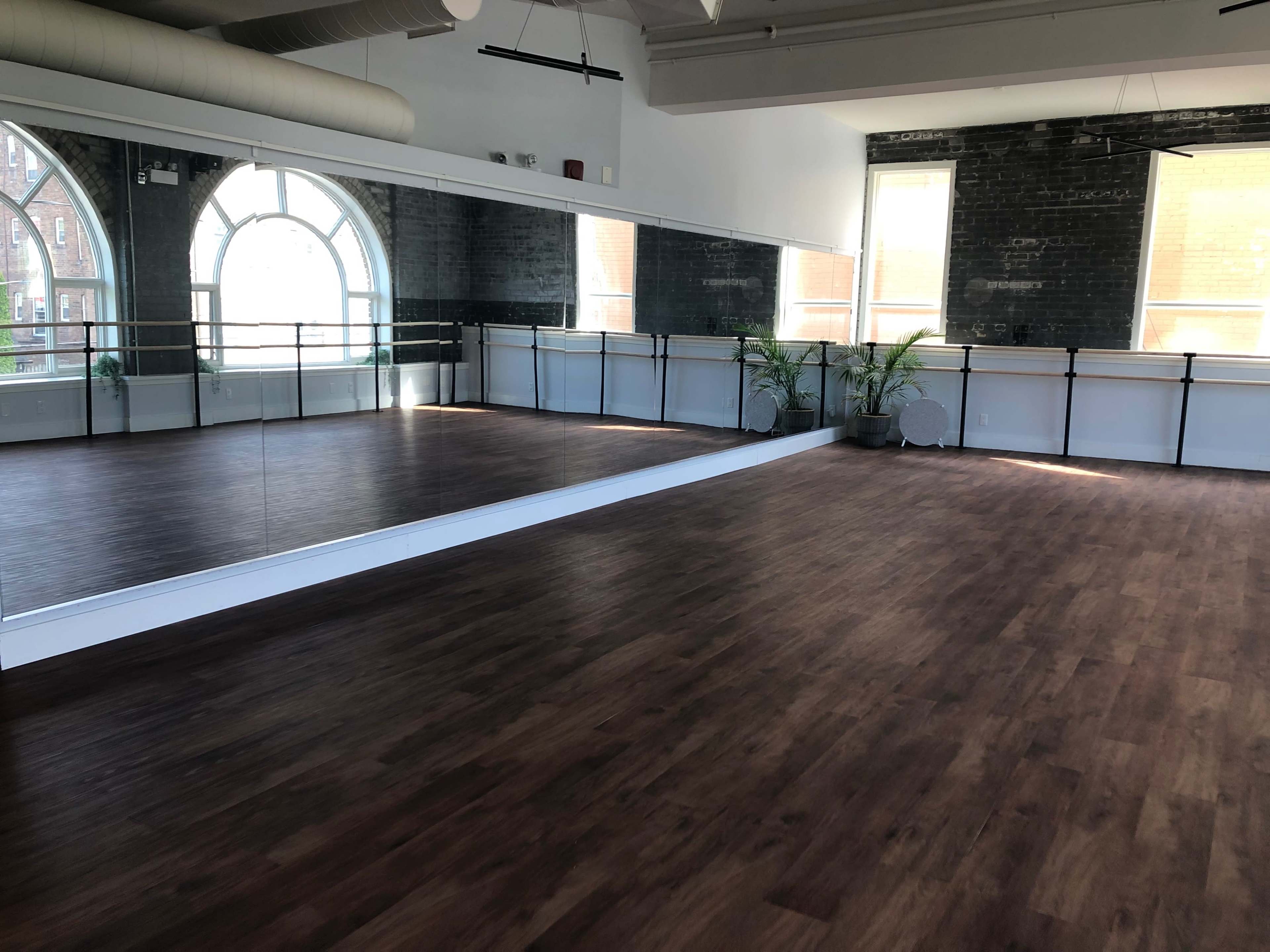 The image shows a spacious dance studio with wooden floors, large mirrors, and windows allowing natural light to enter.