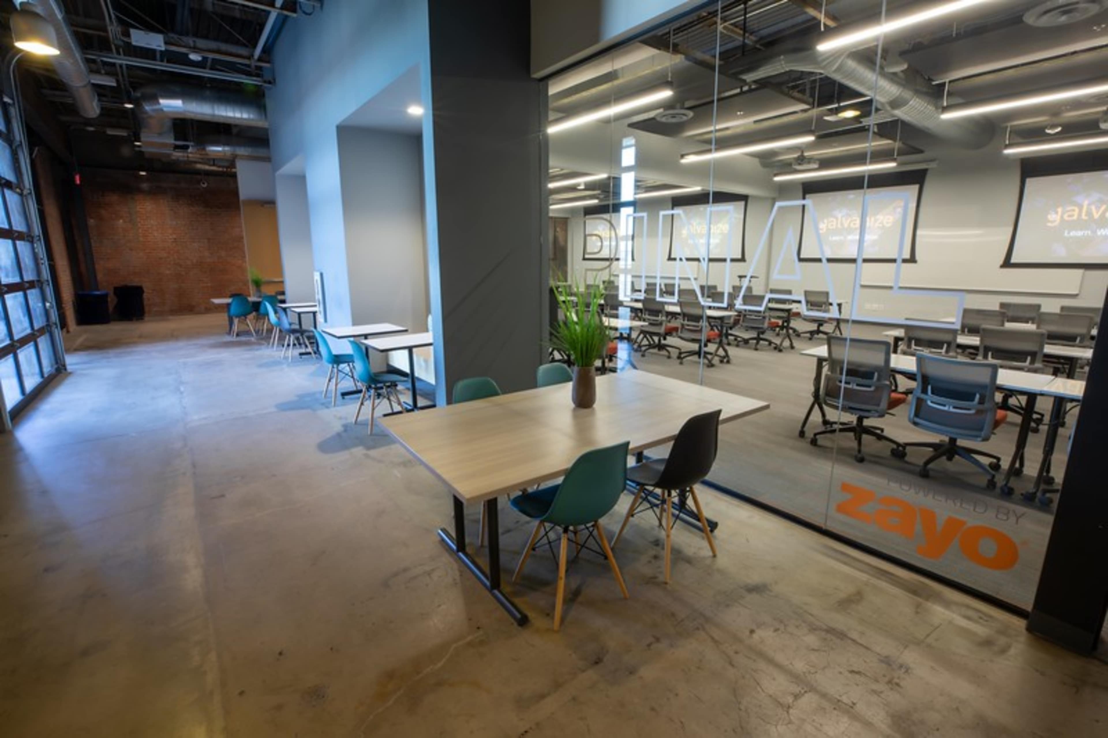 A modern office space with tables and chairs arranged in a well-lit area, alongside a glass-walled meeting room featuring a presentation setup.