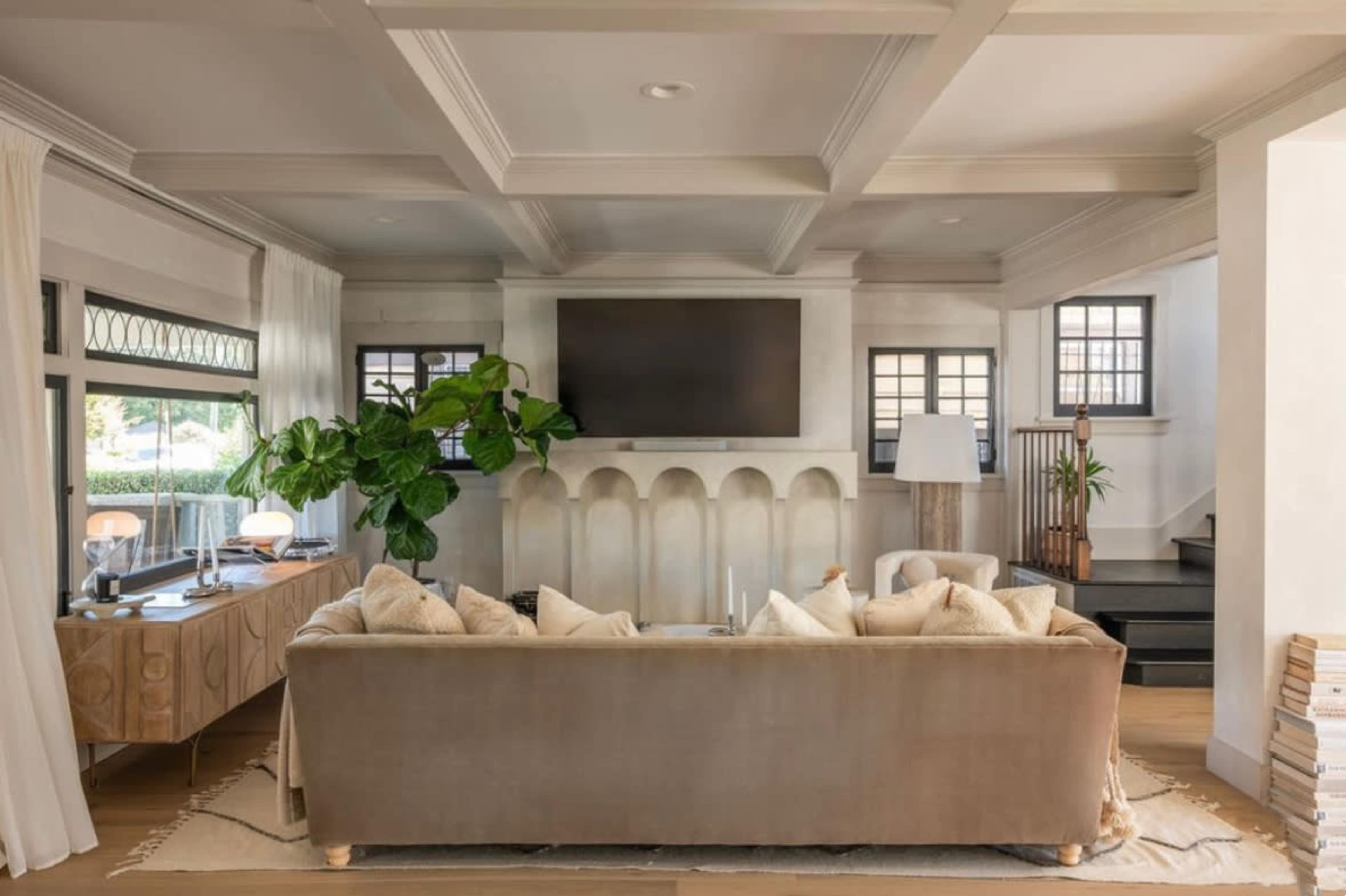The image shows a modern living room featuring a beige sofa, a large television mounted on the wall, and various plants, with light streaming through windows and a staircase visible in the background.