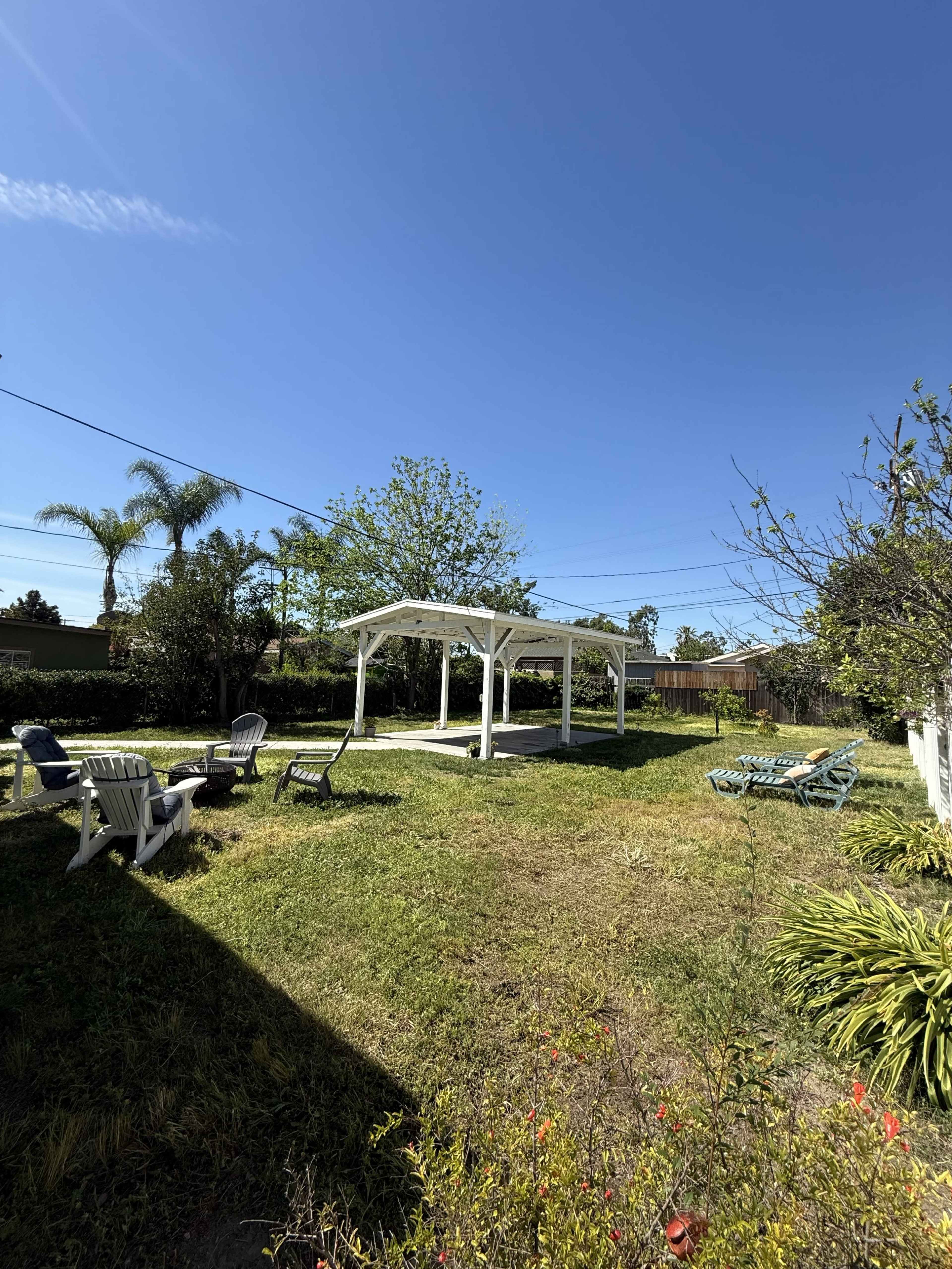 The image shows a grassy yard with a white pergola, surrounded by a few chairs and trees under a clear blue sky.