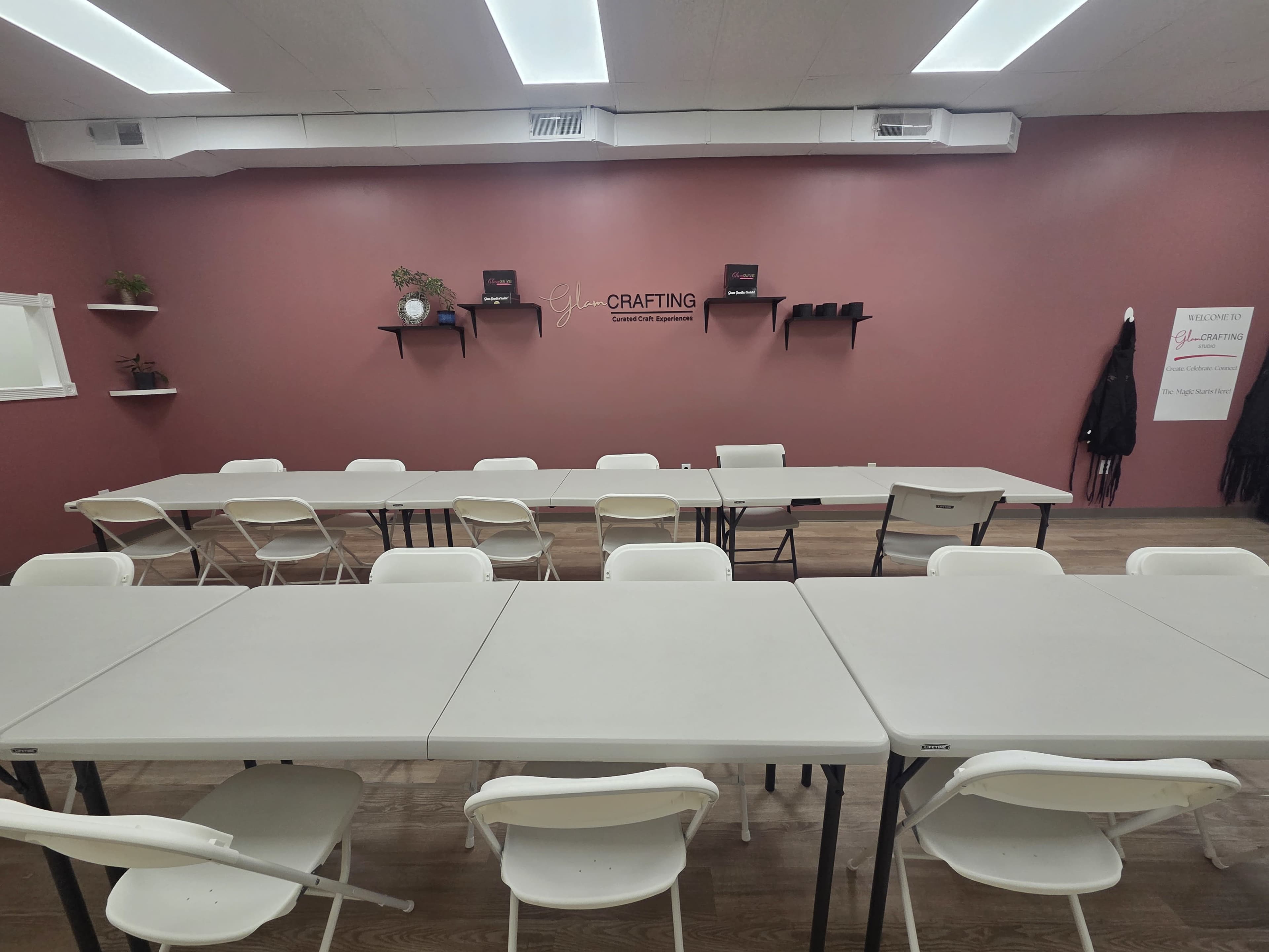 The image shows a spacious room with several tables arranged in a U-shape and white folding chairs, against a pink wall with decorative shelving and a sign.