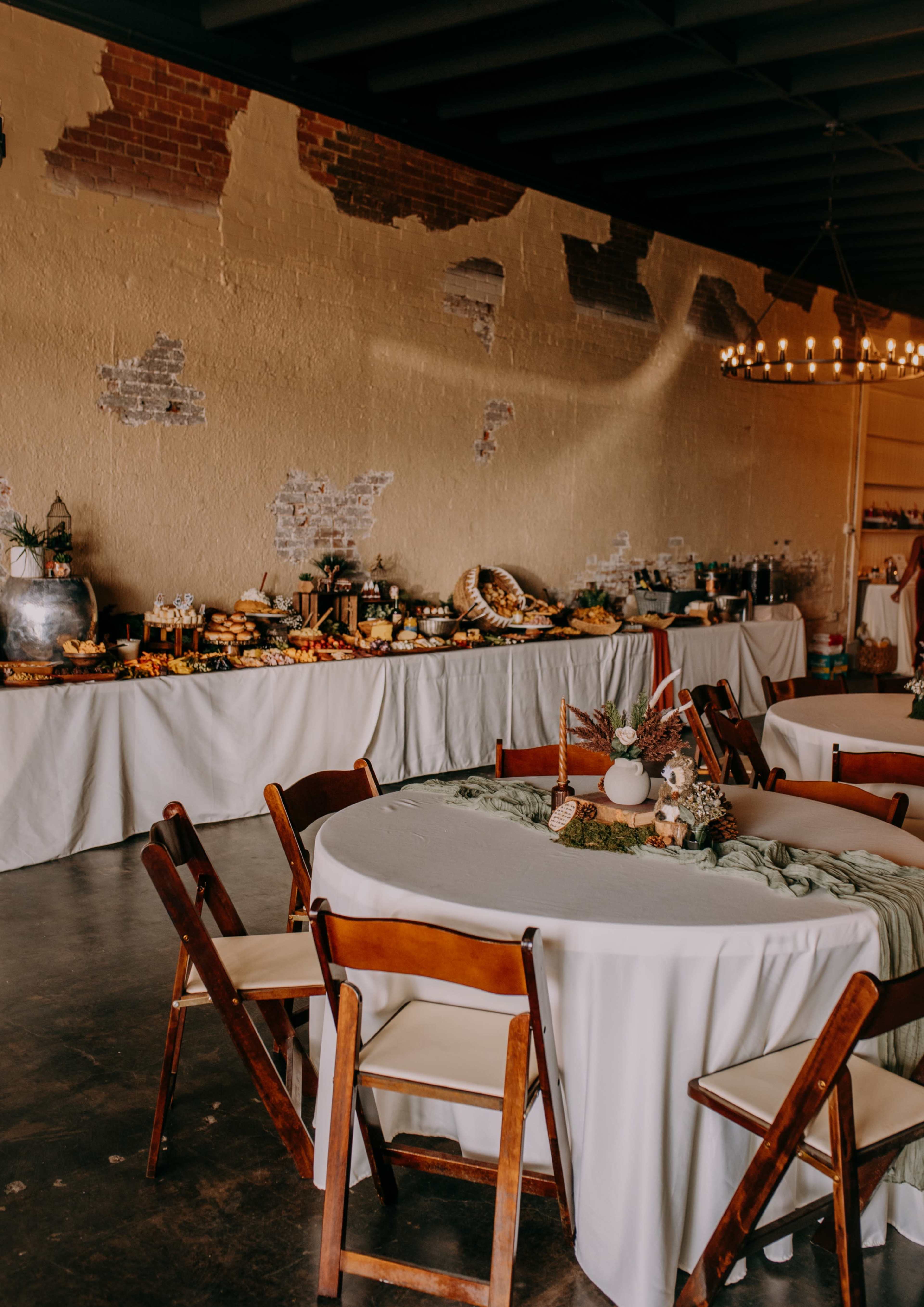 A banquet room features round tables with wooden chairs, and a long buffet filled with various dishes and decorative items along a wall.