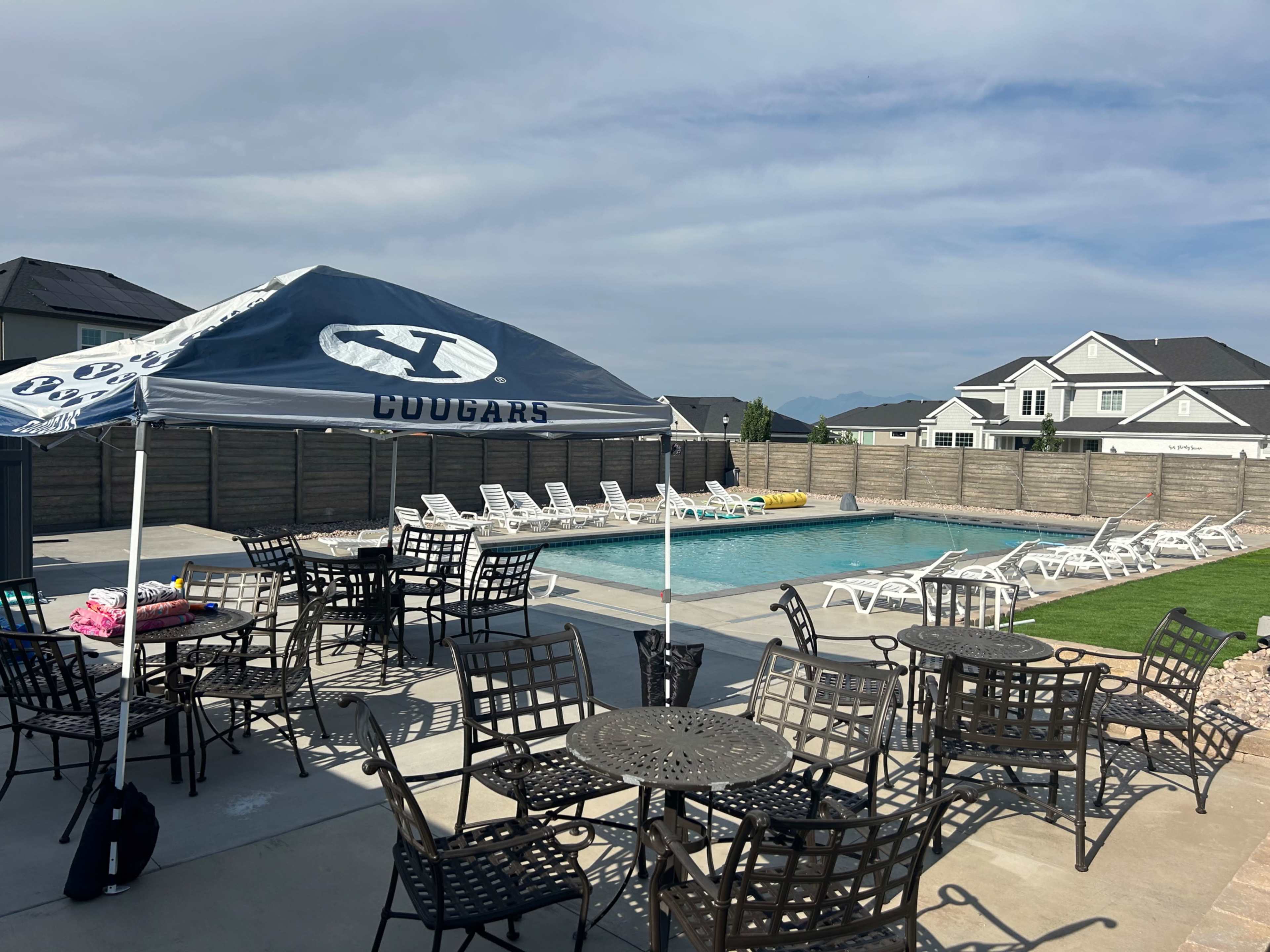 A swimming pool area features a canopy with the word "COUGARS" and is surrounded by tables and lounge chairs, with homes visible in the background.