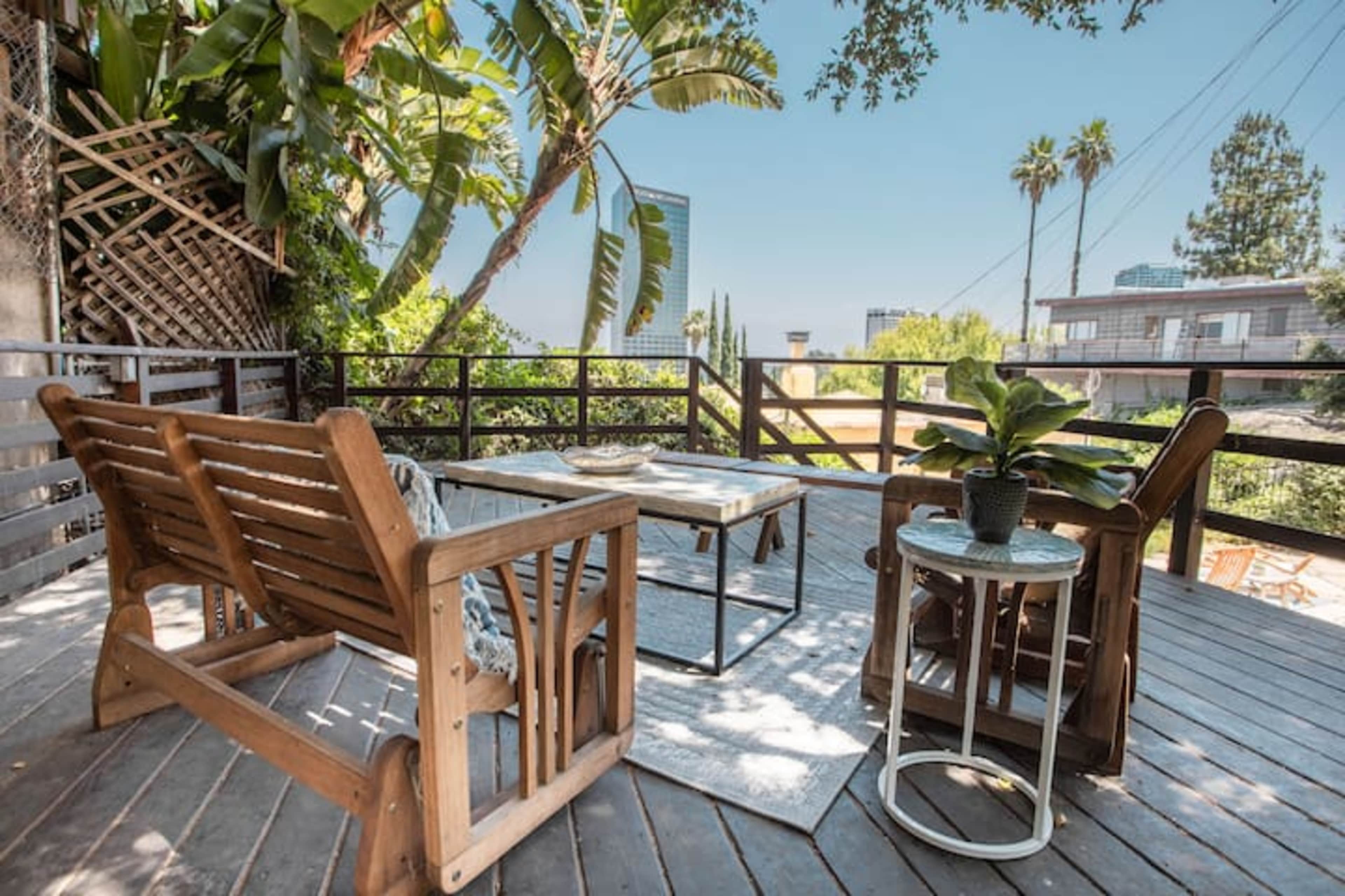 A wooden deck features two chairs and a small table surrounded by tropical plants and a clear blue sky.