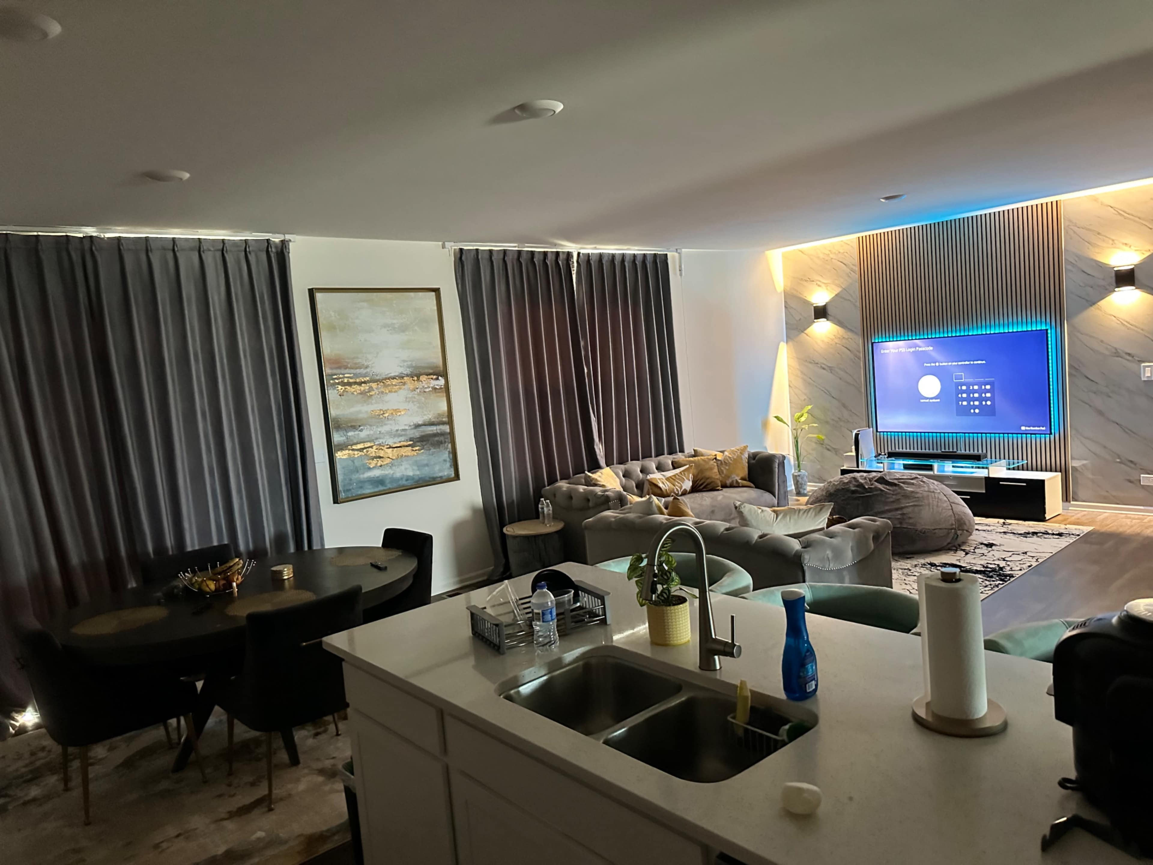 A modern living space with a kitchen in the foreground, a dining area with a black table, and a lounge area featuring a large couch and a wall-mounted TV.