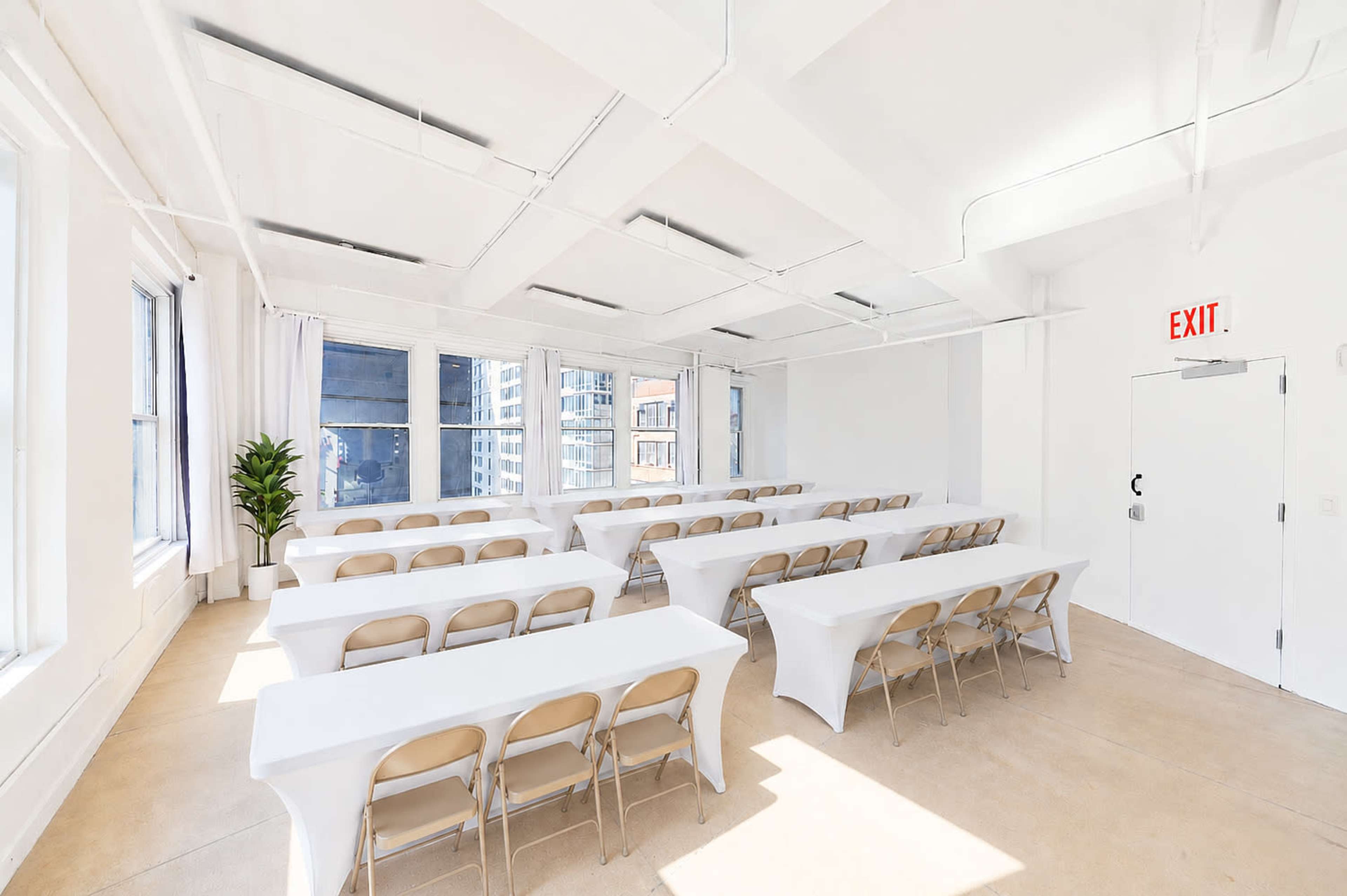 The image shows a bright, empty conference room set up with multiple long tables covered in white cloth and folding chairs arranged in rows, with large windows allowing natural light.
