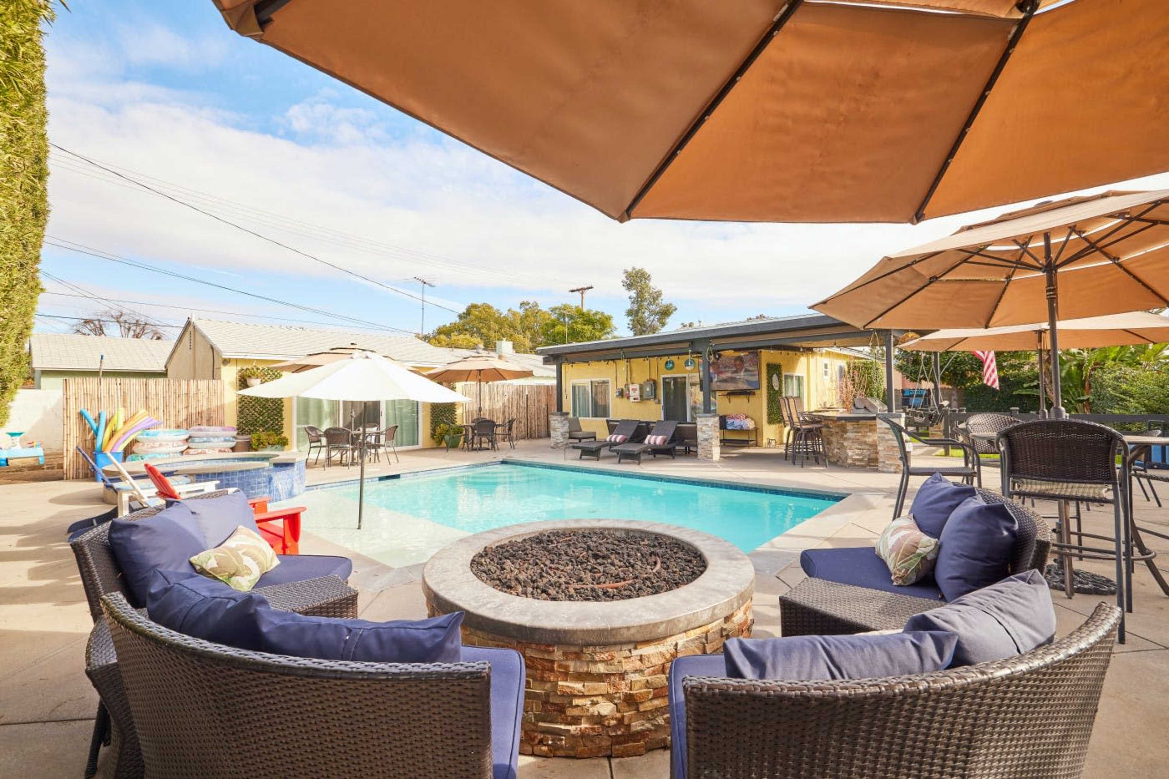 The image shows a backyard pool area with a fire pit surrounded by comfortable seating and several umbrellas for shade.