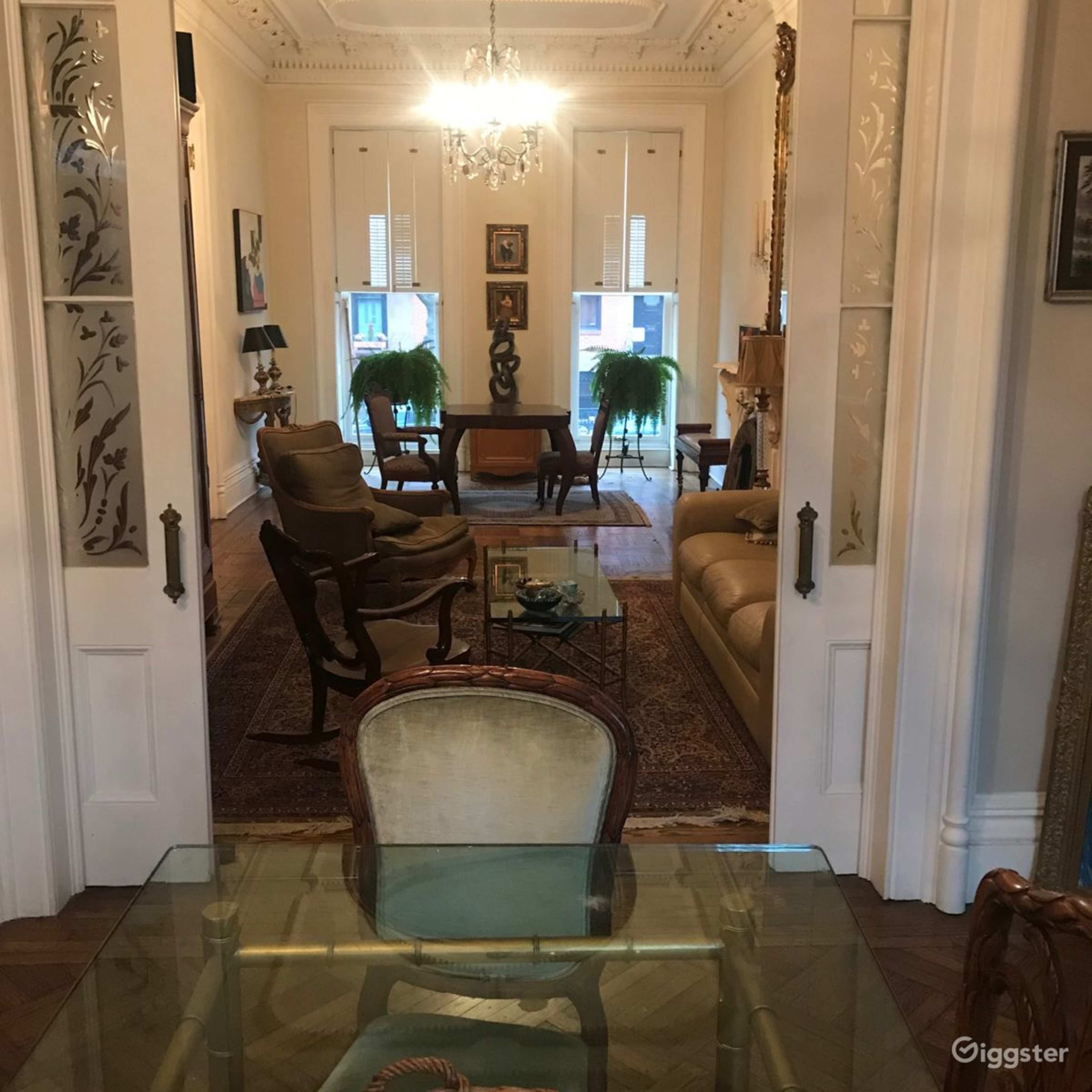 A spacious living room with two large plants, a sofa, armchairs, and a clear view through open doors leading to another area.