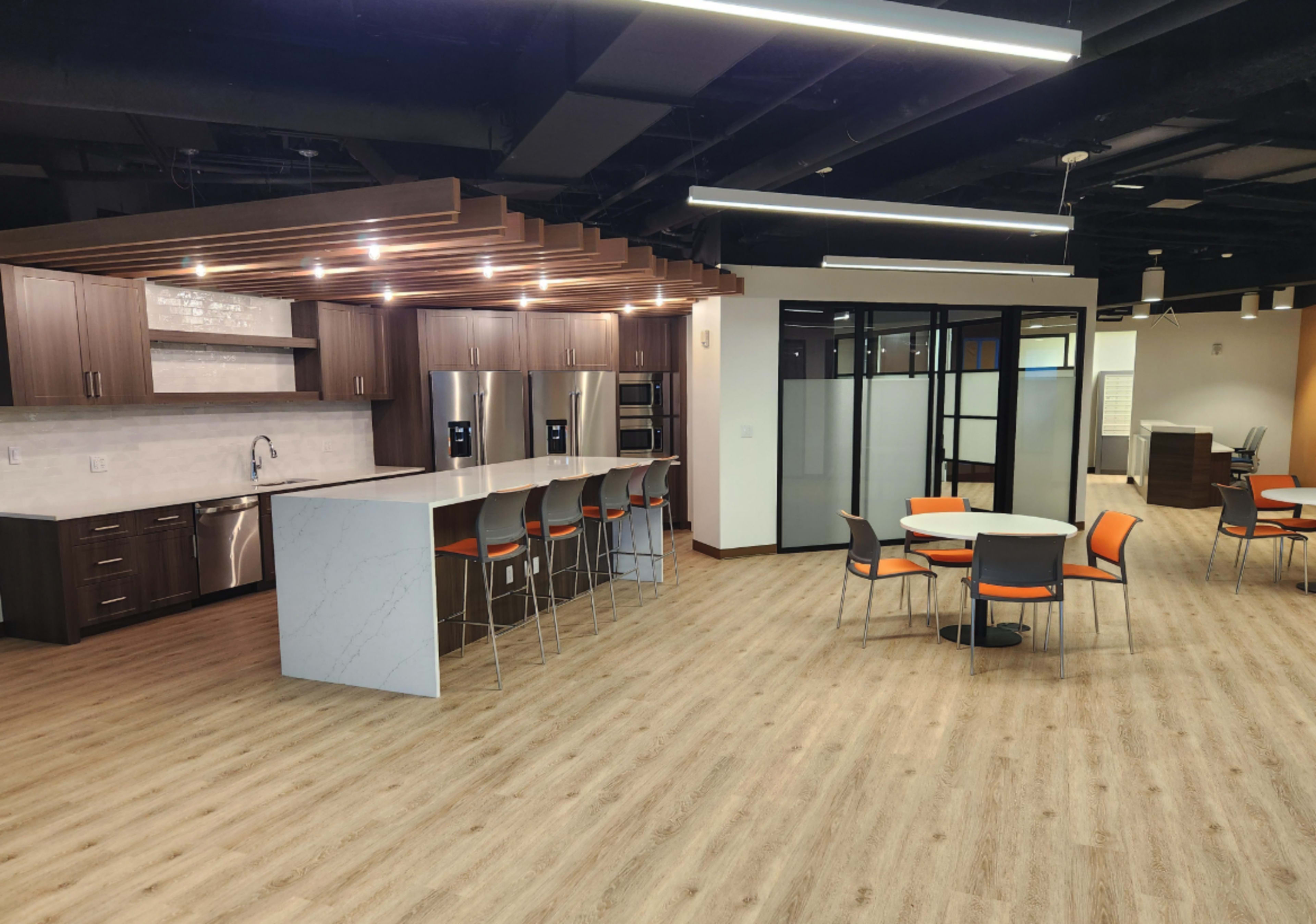 The image shows a modern kitchen and dining area with sleek cabinetry, stainless steel appliances, a large island with bar seating, and a spacious seating arrangement featuring round tables and chairs.