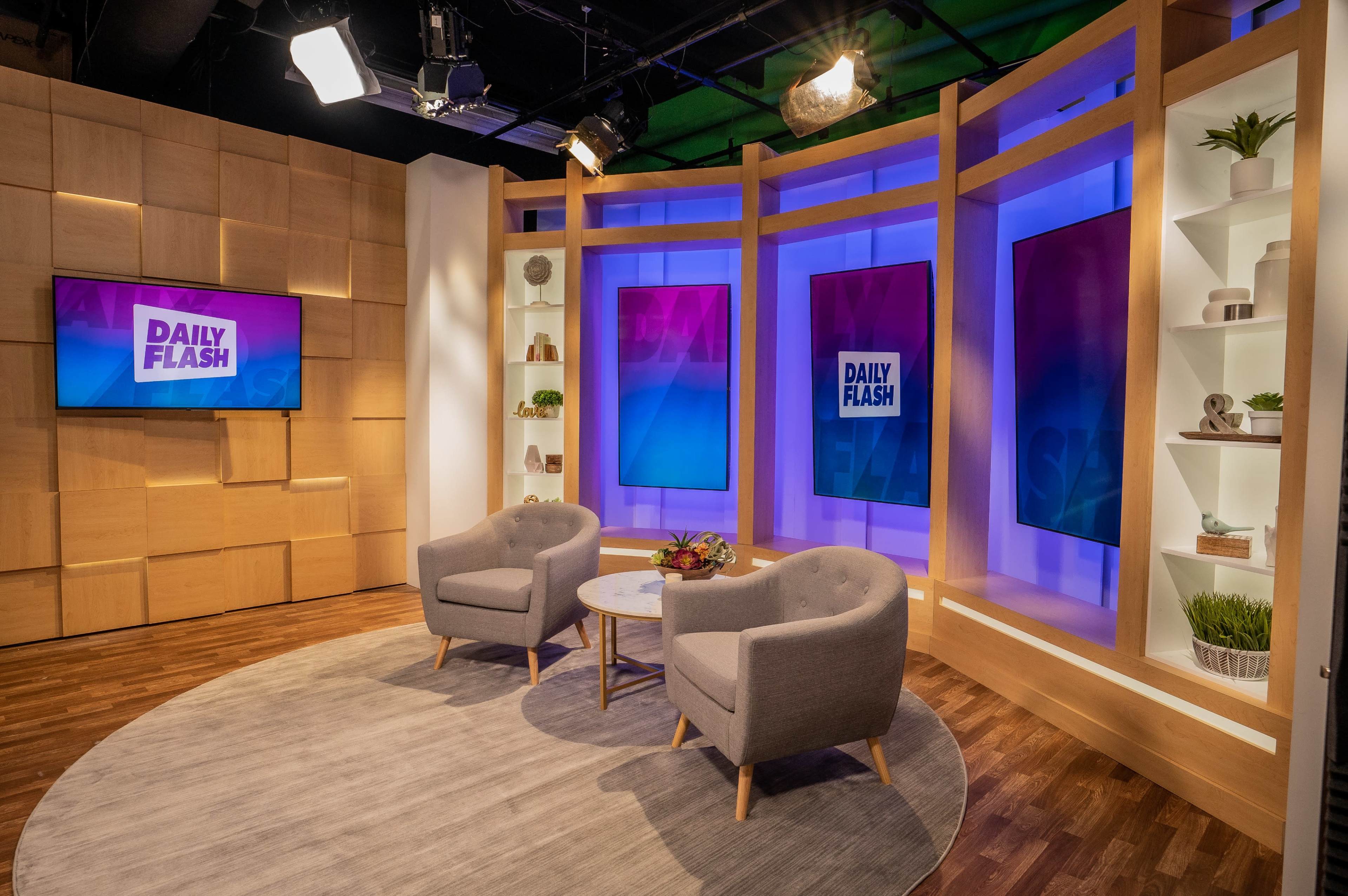 The image shows a television news studio set with two gray chairs and a round table, illuminated by colorful backdrops and a screen displaying "DAILY FLASH."