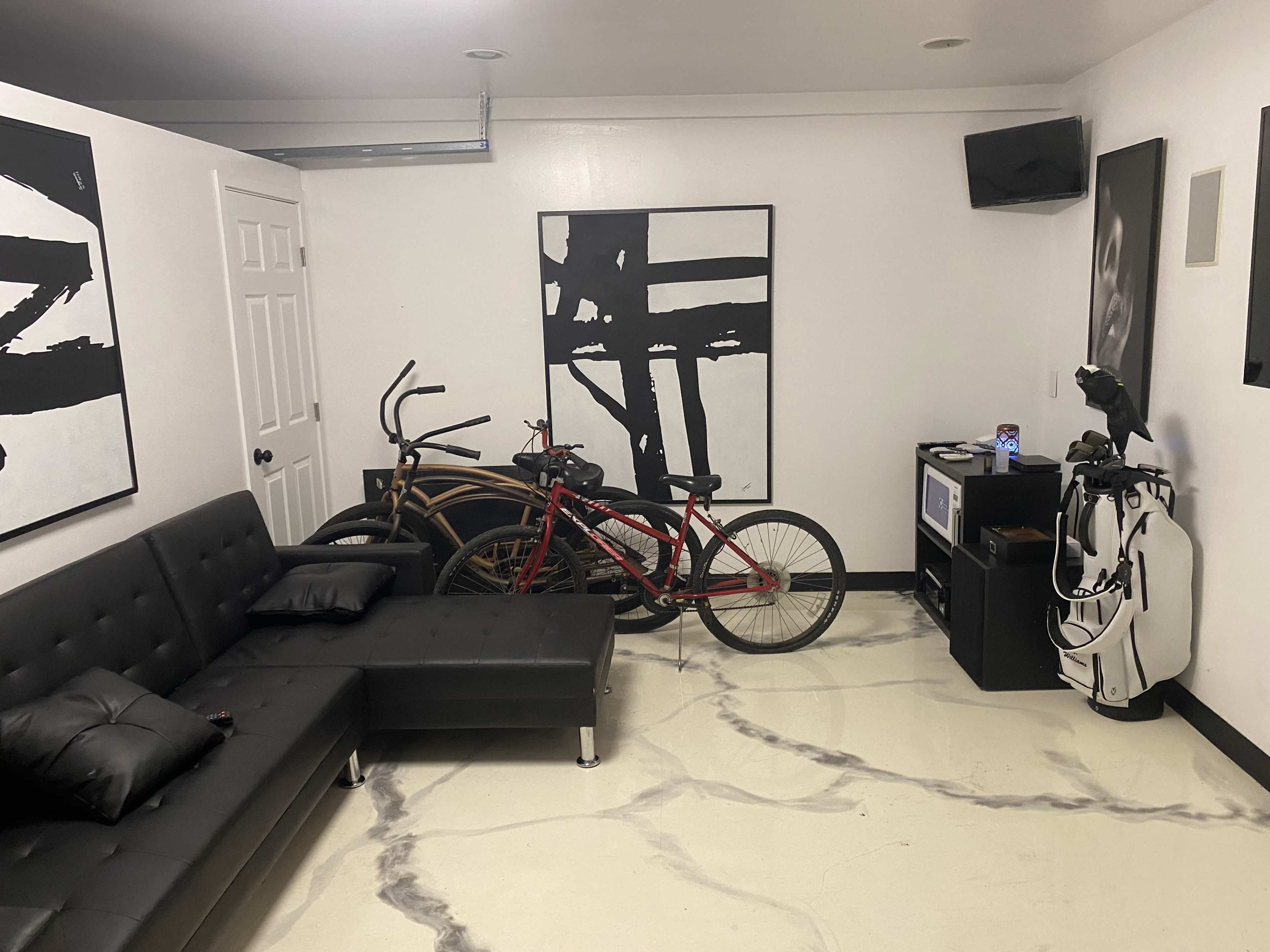 A room featuring two bicycles parked next to a black couch, a television, and a small cabinet in a space with white walls and a marbled floor.
