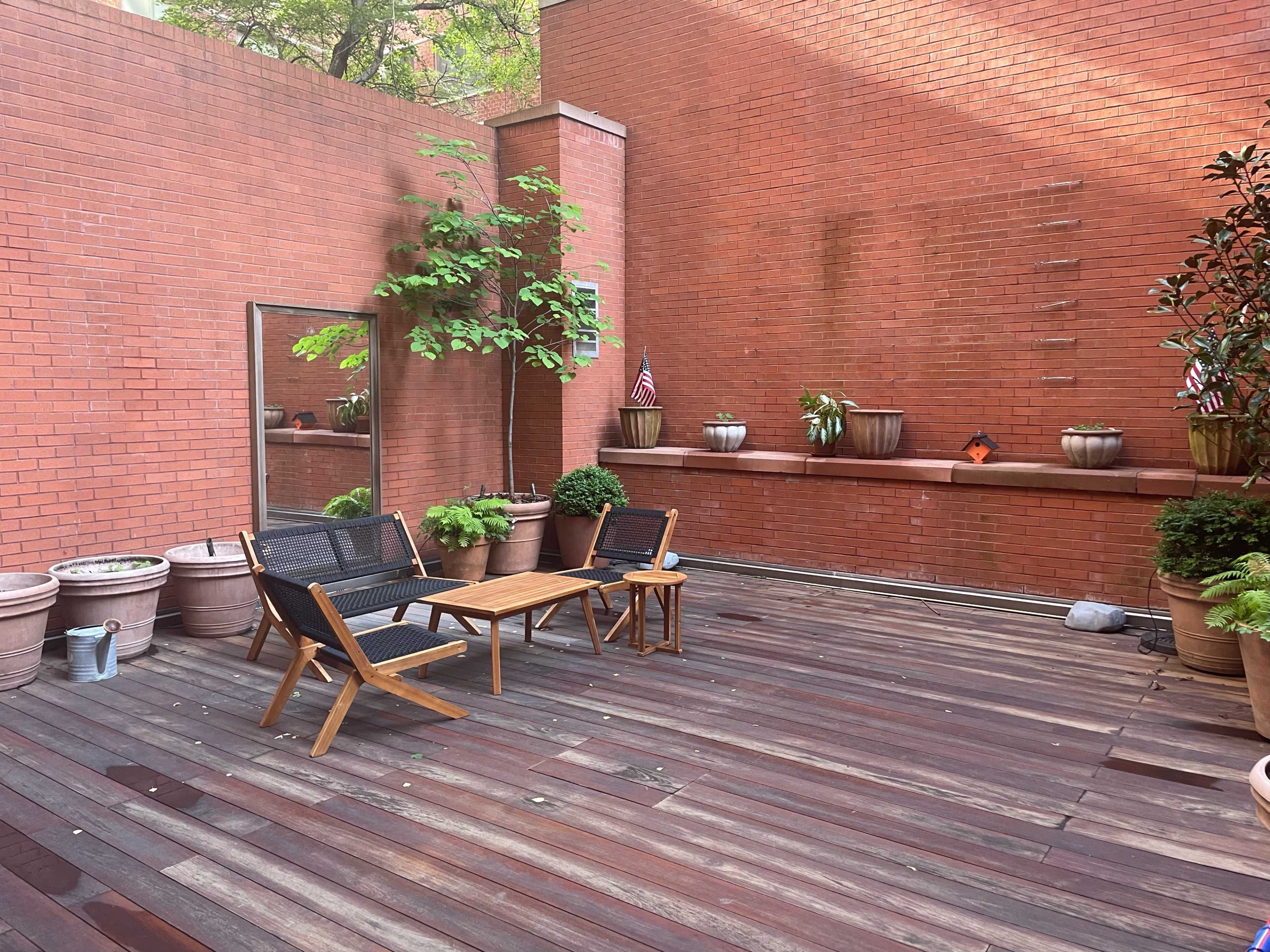 The image shows a wooden patio area with two chairs and a small table, surrounded by potted plants and a mirror against a red brick wall.