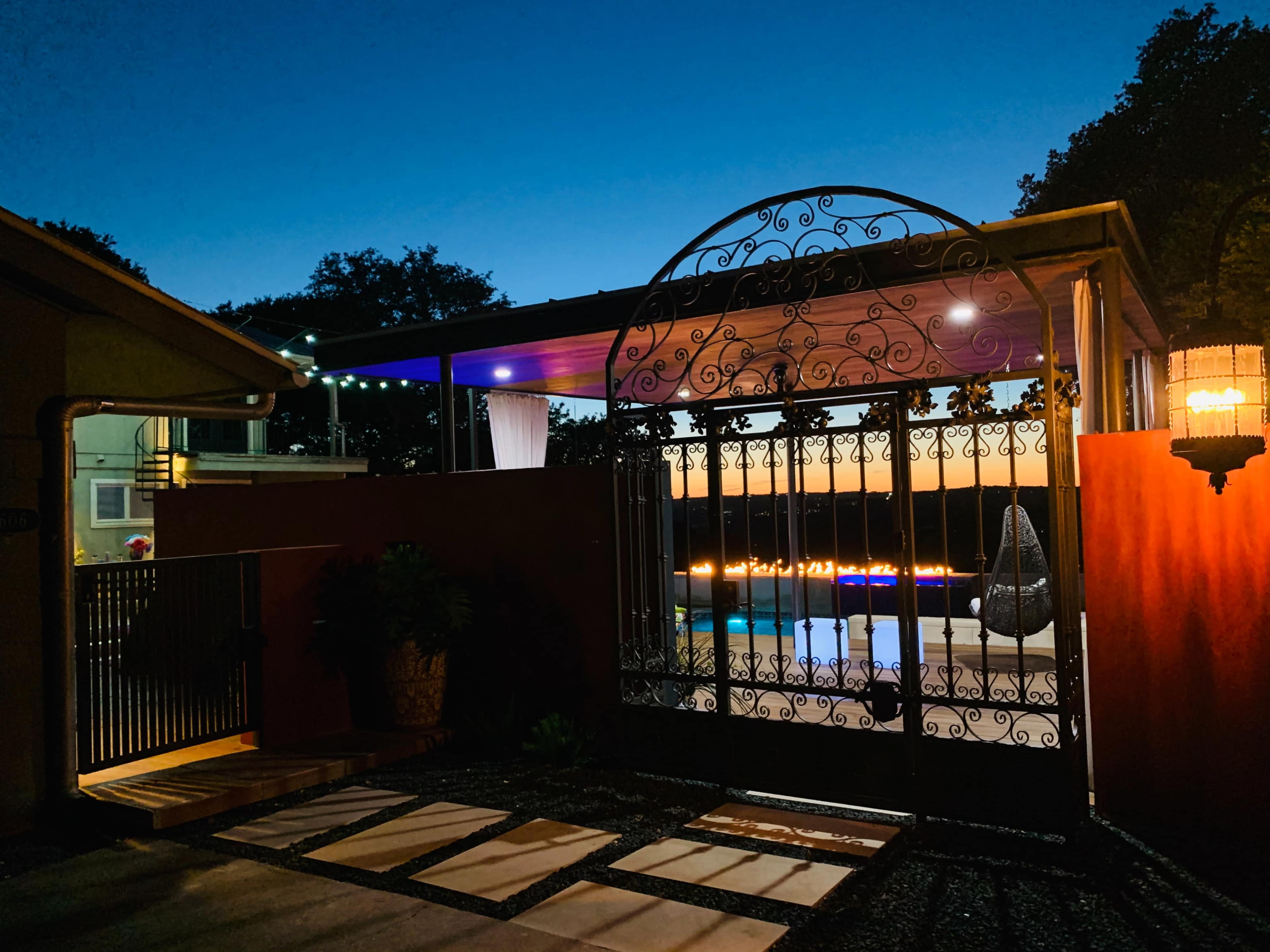 A wrought iron gate opens to a well-lit patio with a pool, surrounded by decorative plants and accent lighting at dusk.